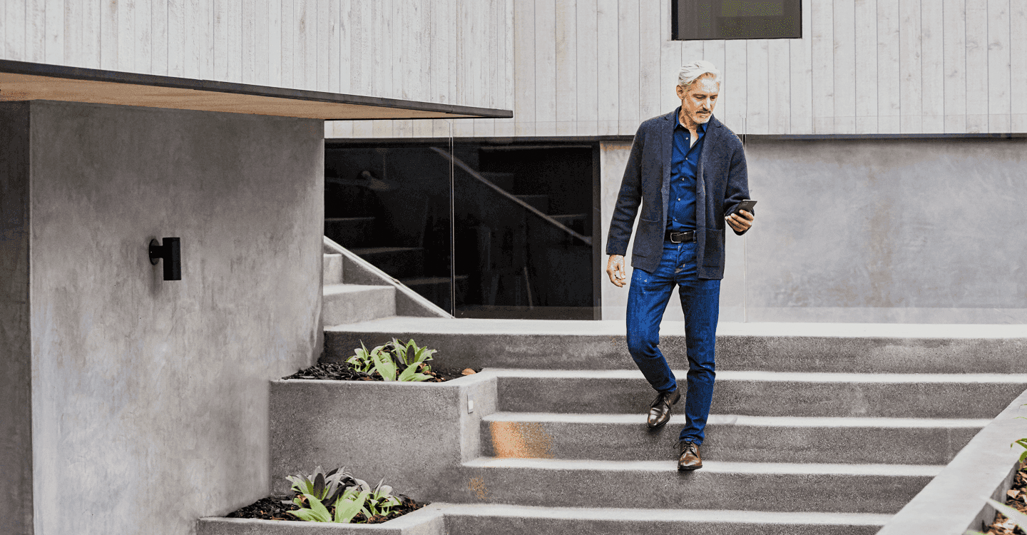 An older man with gray hair, dressed in a navy blazer, blue shirt, and jeans, walking down outdoor concrete stairs while looking at his smartphone.
