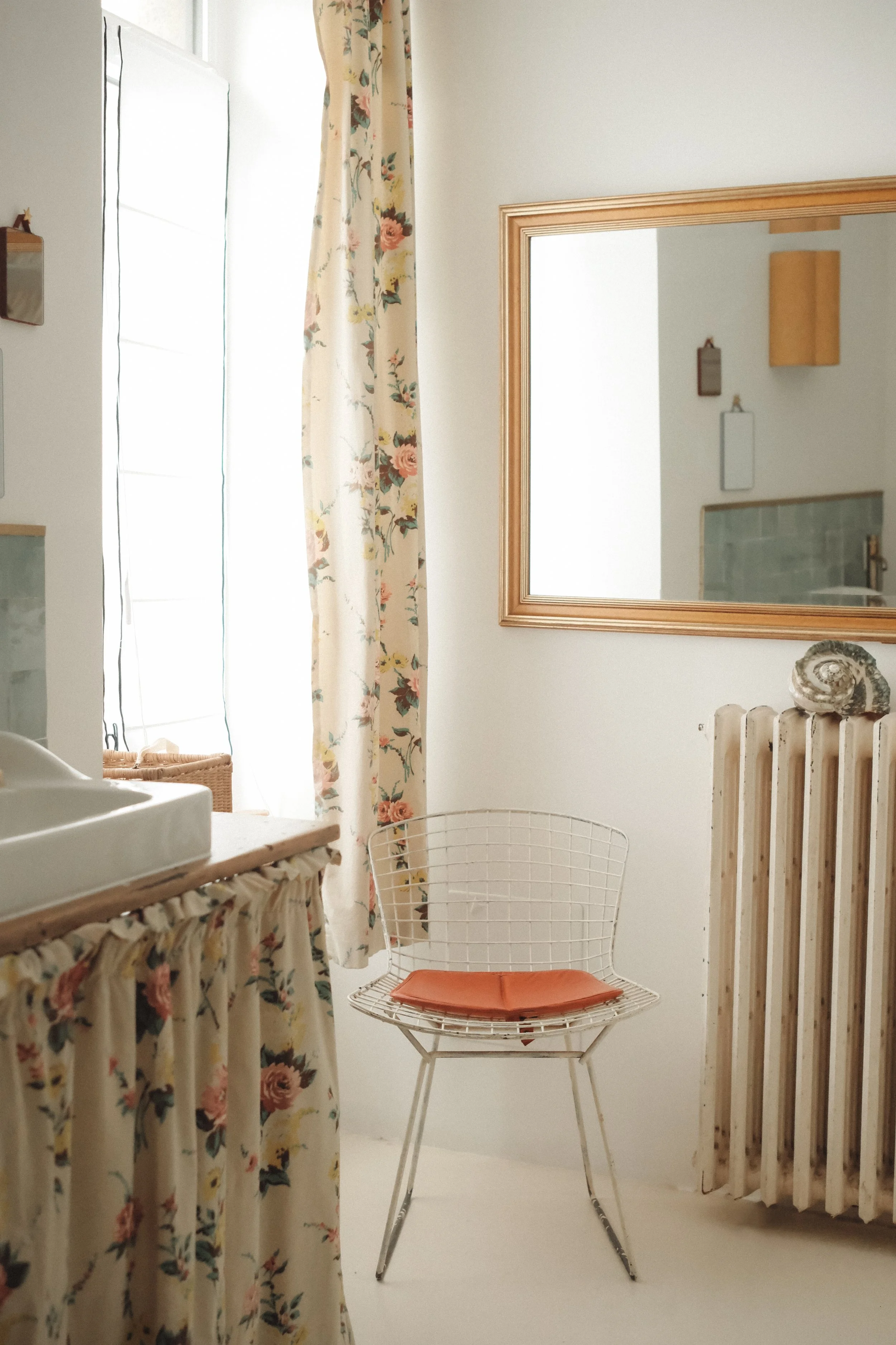 Room with floral curtain, wicker basket, mirror, radiator with seashell on top, white wire chair with orange cushion, and part of a sink.
