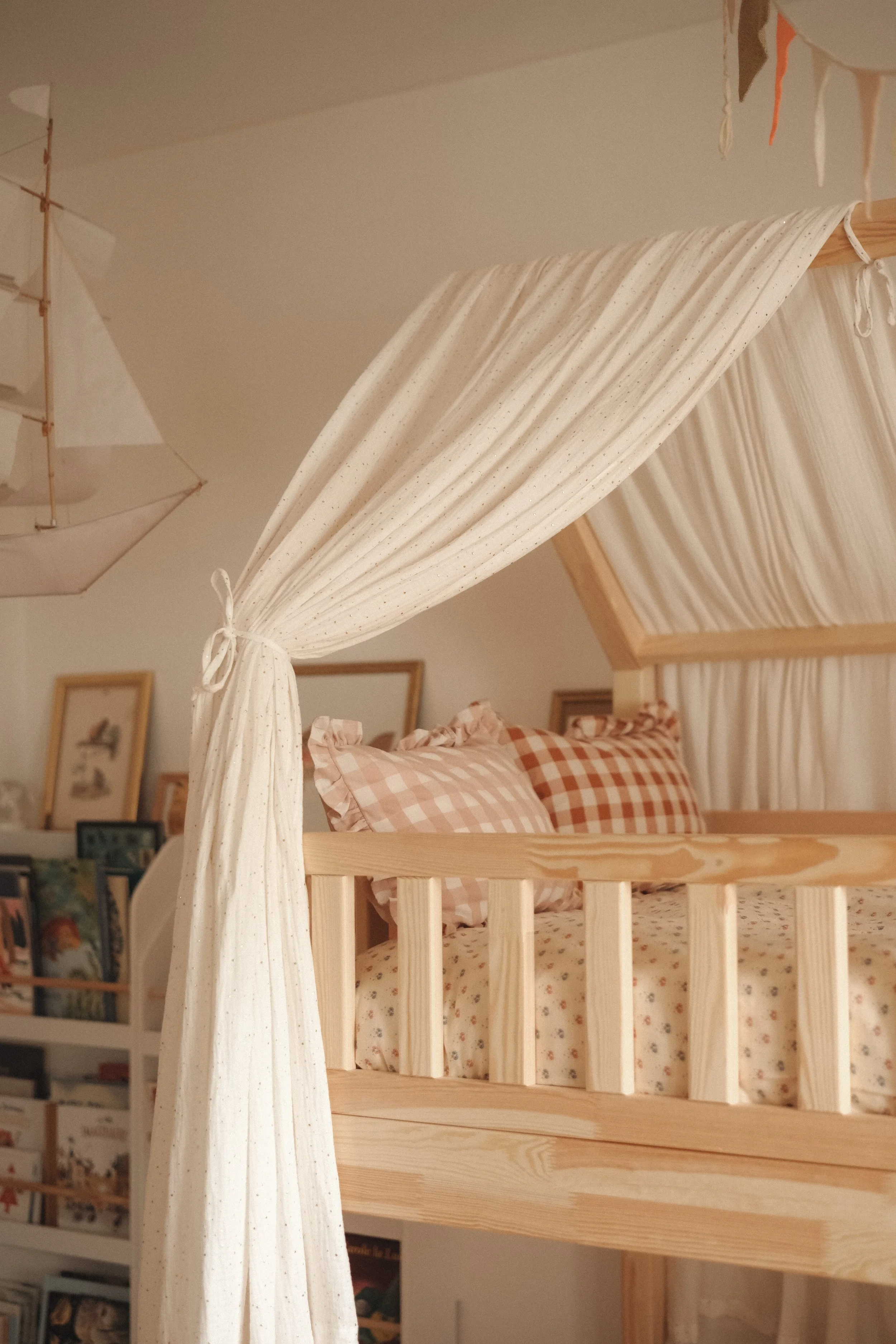 A child's bedroom with a wooden canopy bed and plaid pillows. There is a bookshelf with books and framed pictures in the background. A fabric canopy is draped around the bed.