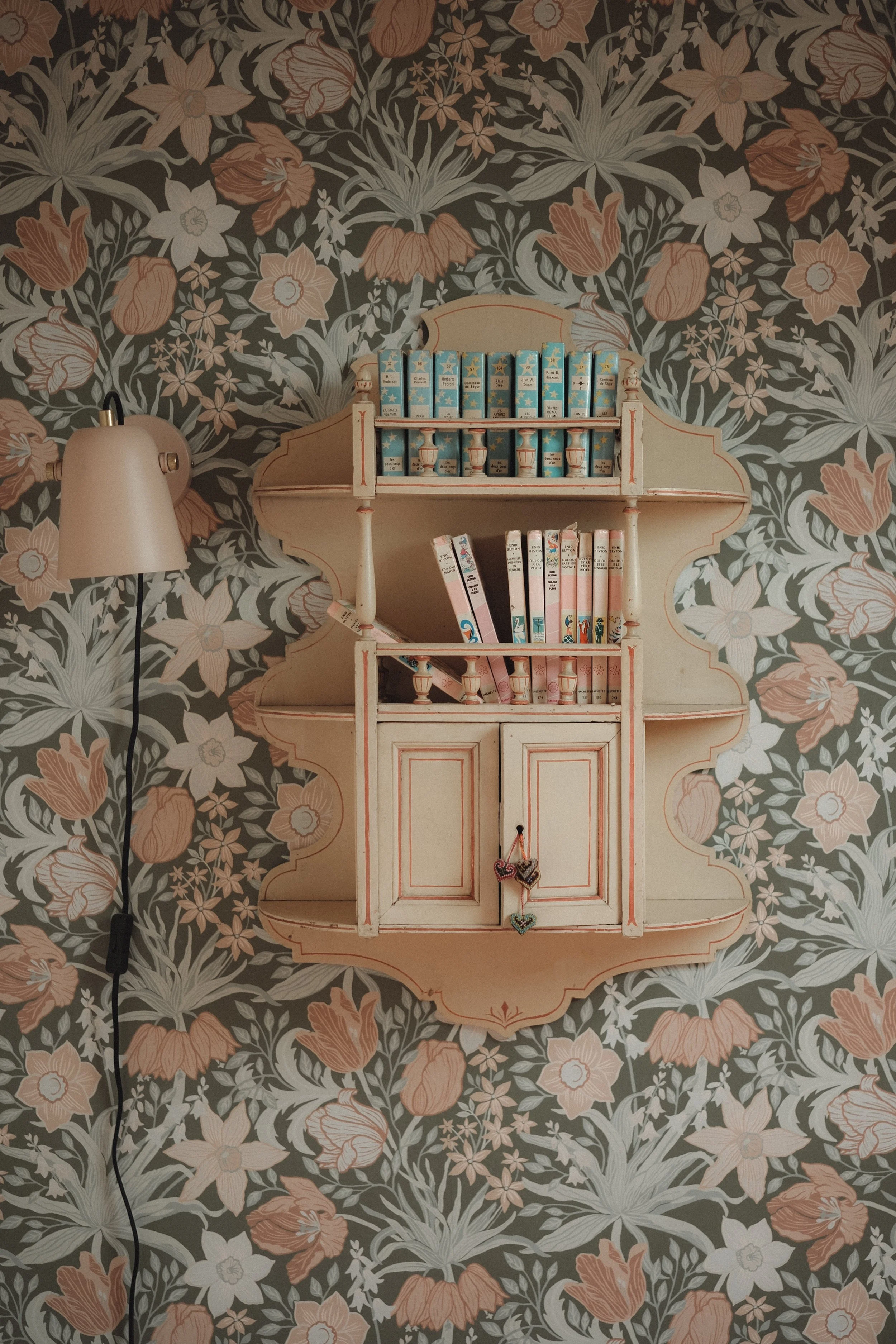 A vintage-style wall shelf with multiple compartments, decorated with books and small ornate items, mounted on a floral wallpaper background with a floor lamp to the left.