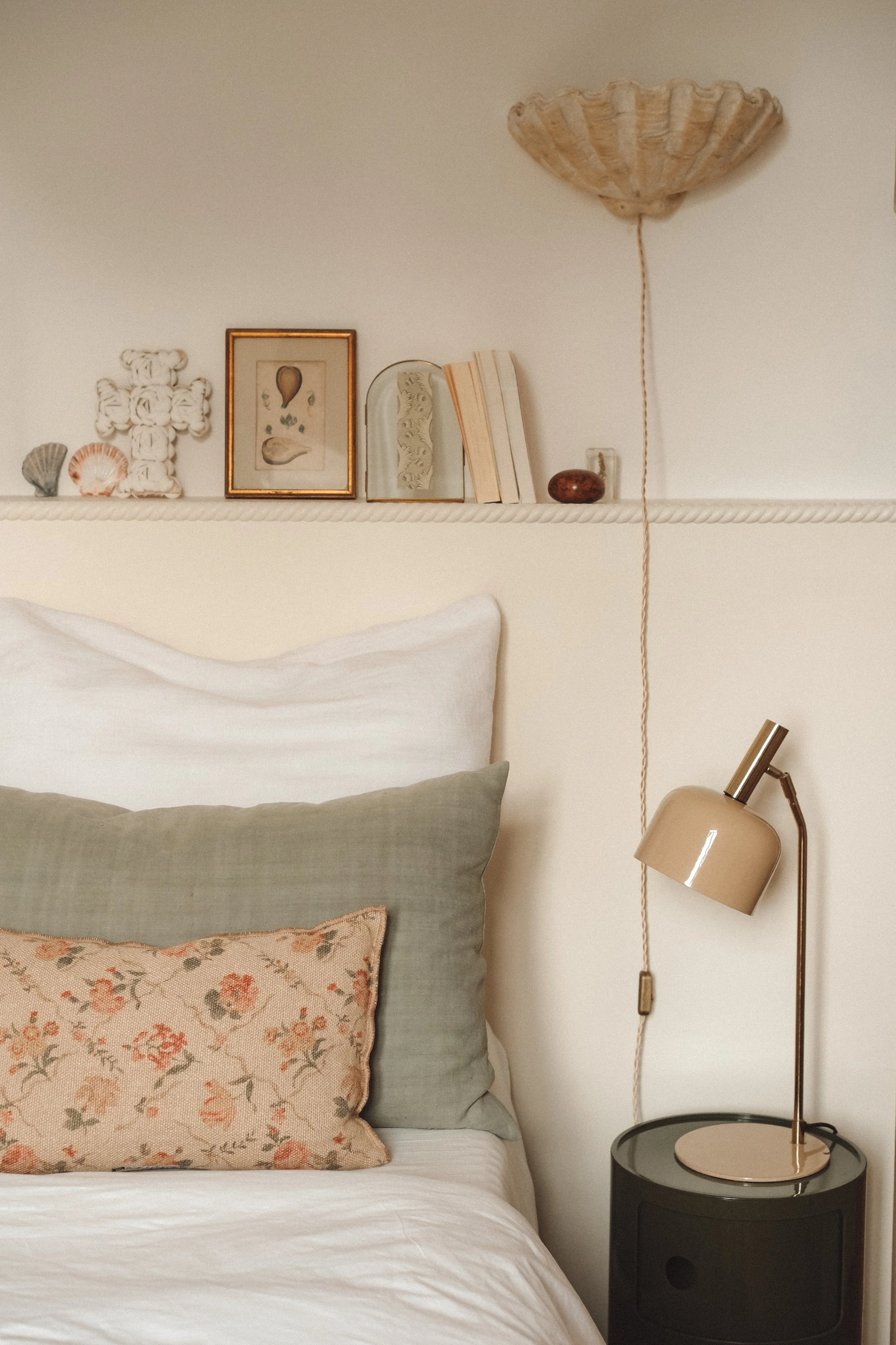 A cozy bedroom featuring a bed with petted white, green, and floral pillows, a black bedside table with a beige desk lamp, and a decorative shelf with books, framed art, seashells, and small decorative items. A textured off-white lampshade hangs from