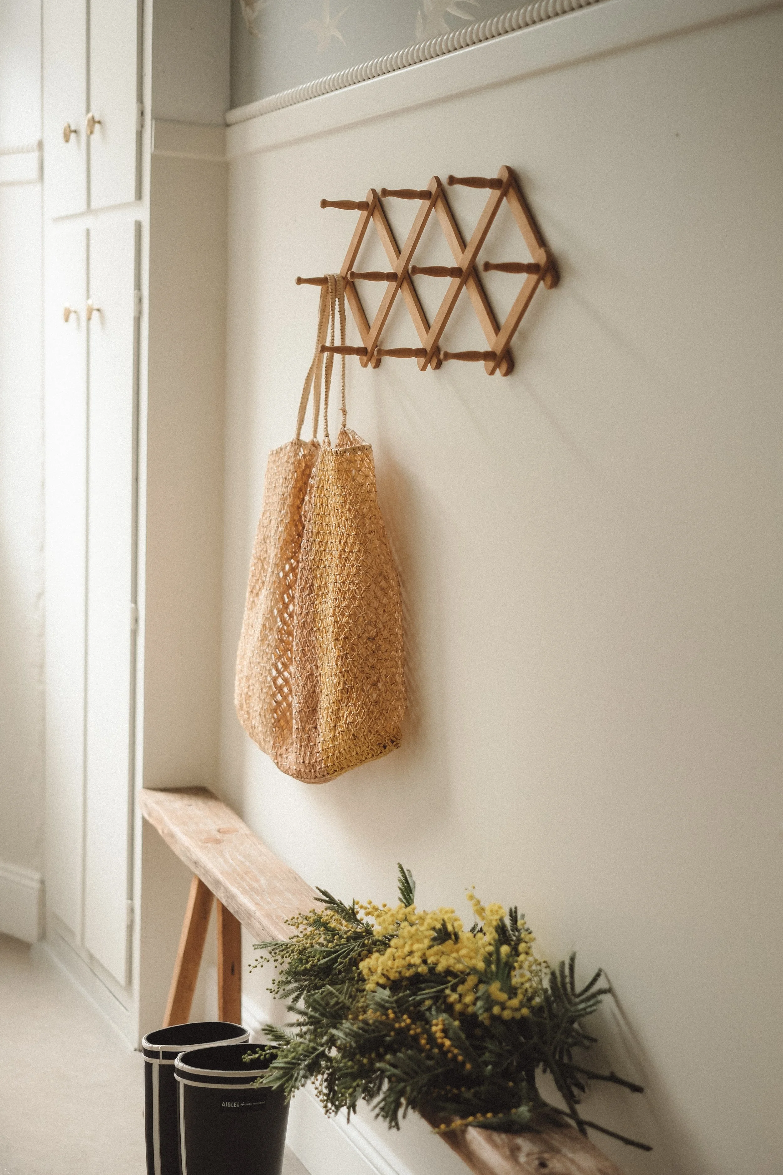 A beige woven bag hanging on a wall hook in a room with white walls, a wooden bench below it with a bunch of yellow flowers on top, and black rain boots nearby. There are also built-in white cabinets to the side.
