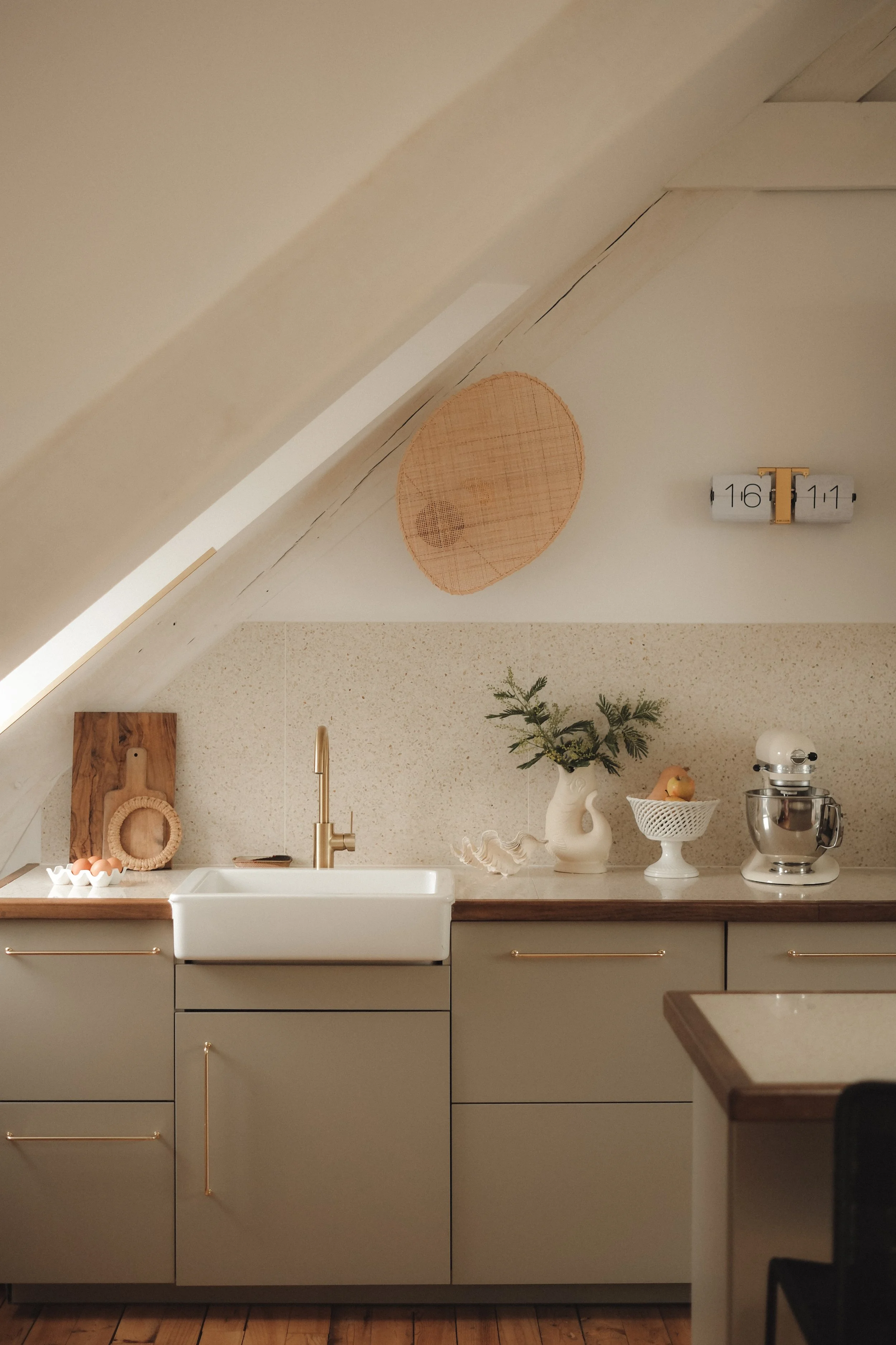 A cozy kitchen with a slanted ceiling, white cabinetry with gold handles, a large white farmhouse sink with a gold faucet, decorative items like a vase with greenery, a fruit bowl, a stand mixer, and a wood cutting board with eggs. There are also wal