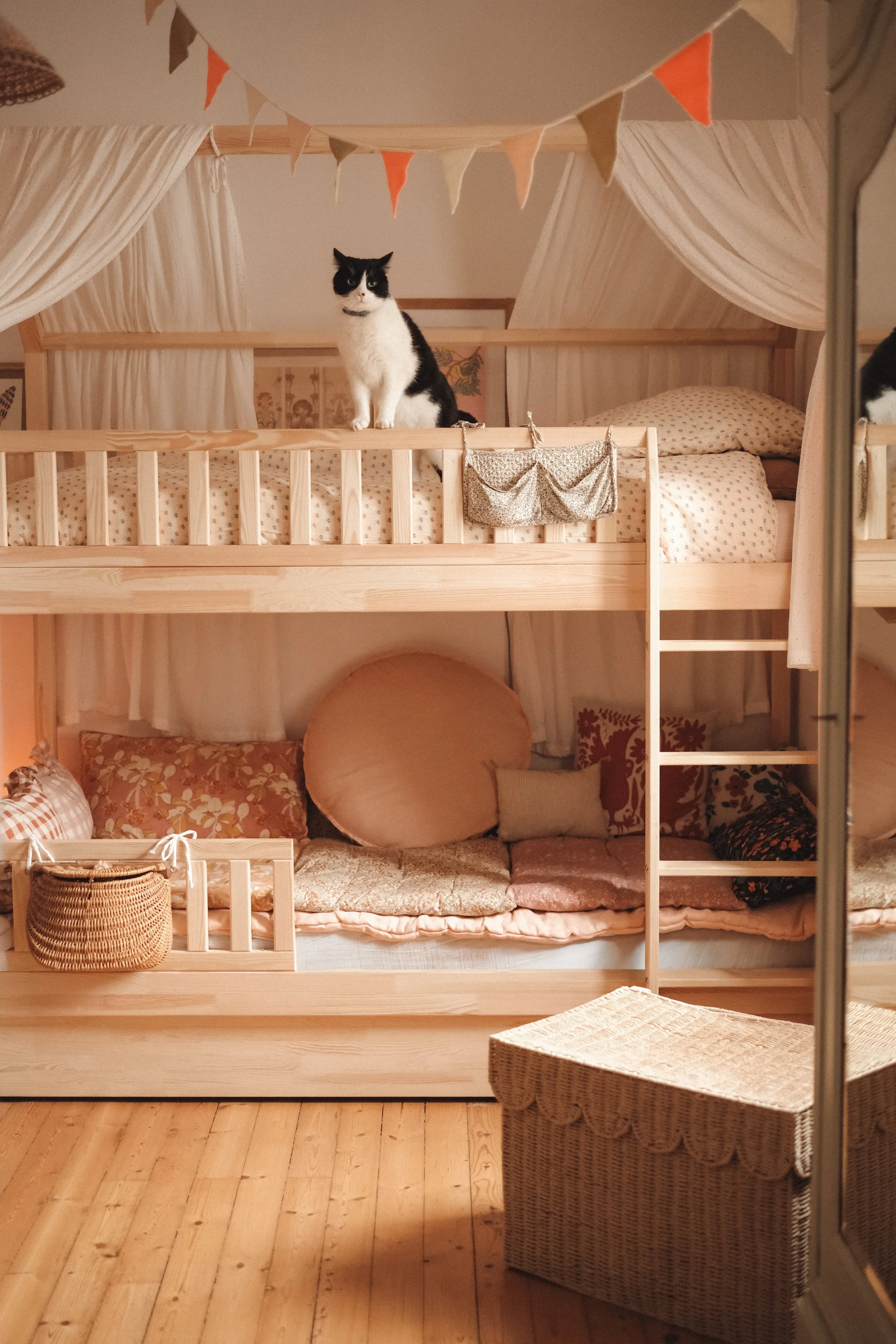 A black and white cat sitting on the top bunk of a wooden bed with a canopy in a cozy room decorated with pastel and floral pillows, cushions, and a hanging bunting banner.