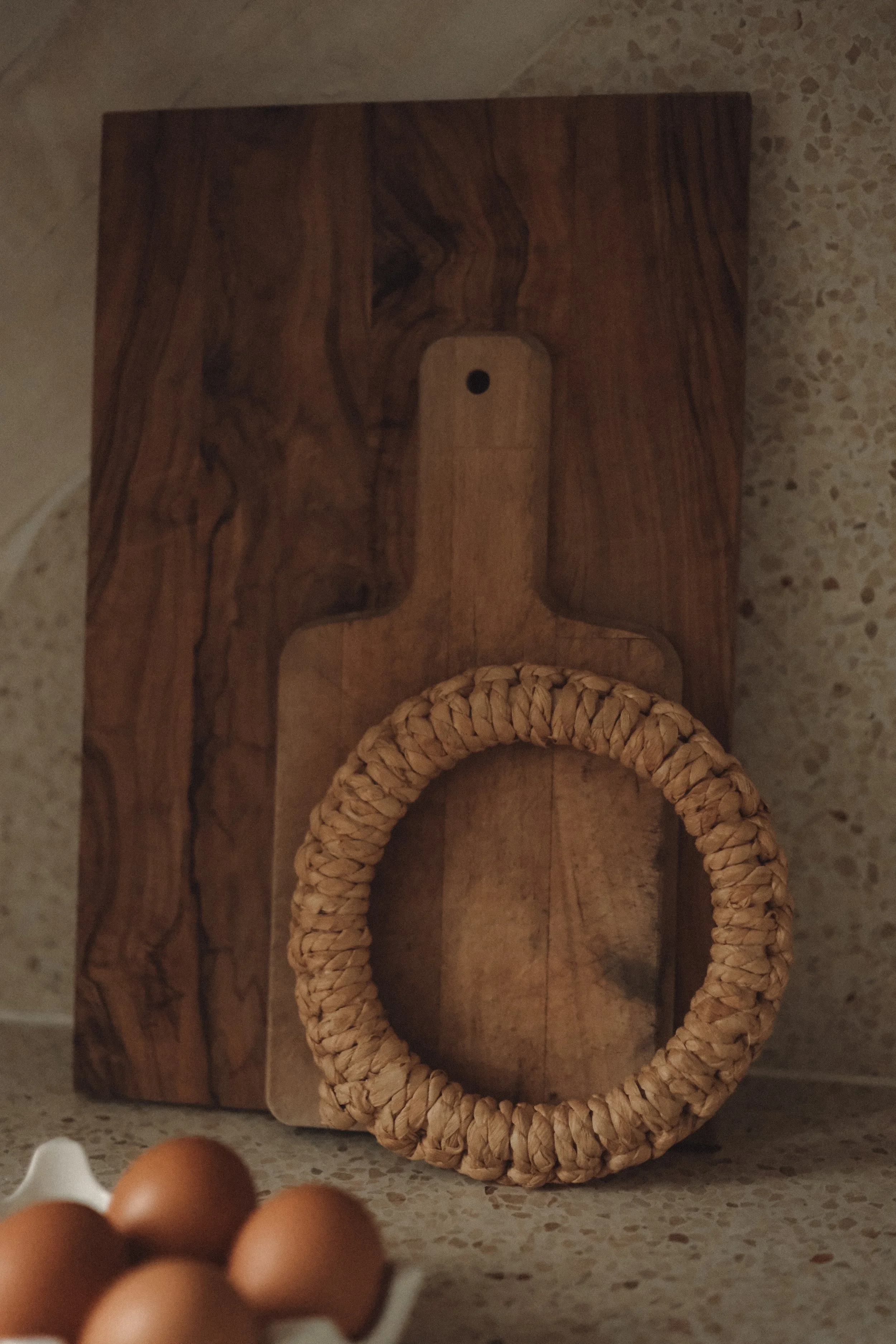 A wooden cutting board with a handle, a round woven straw handle, and a few eggs in a small container on a speckled countertop.