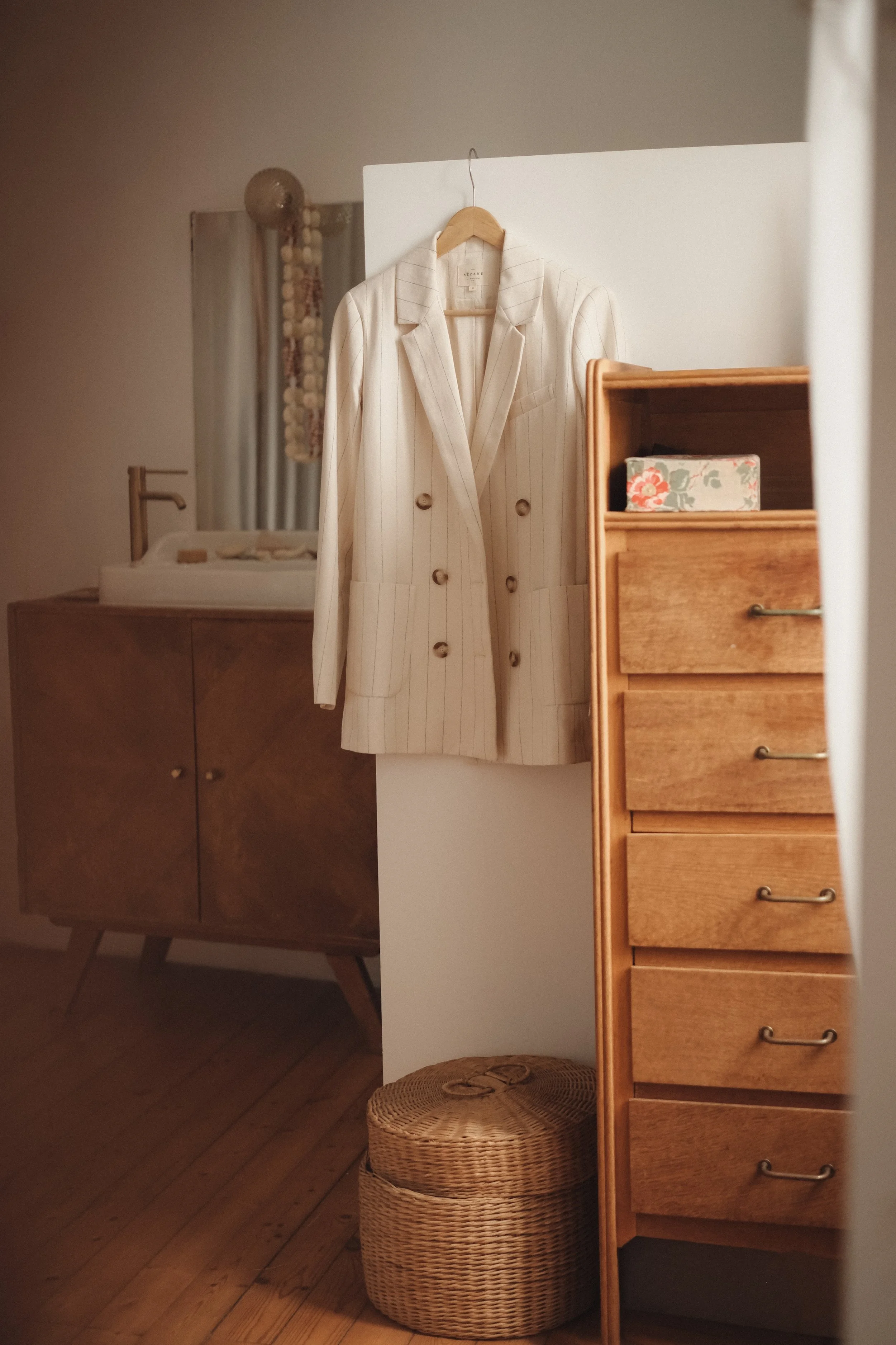 A striped cream blazer hanging on a wooden hanger on a white room divider, with a wooden dresser and wicker basket in a cozy, warmly lit interior.