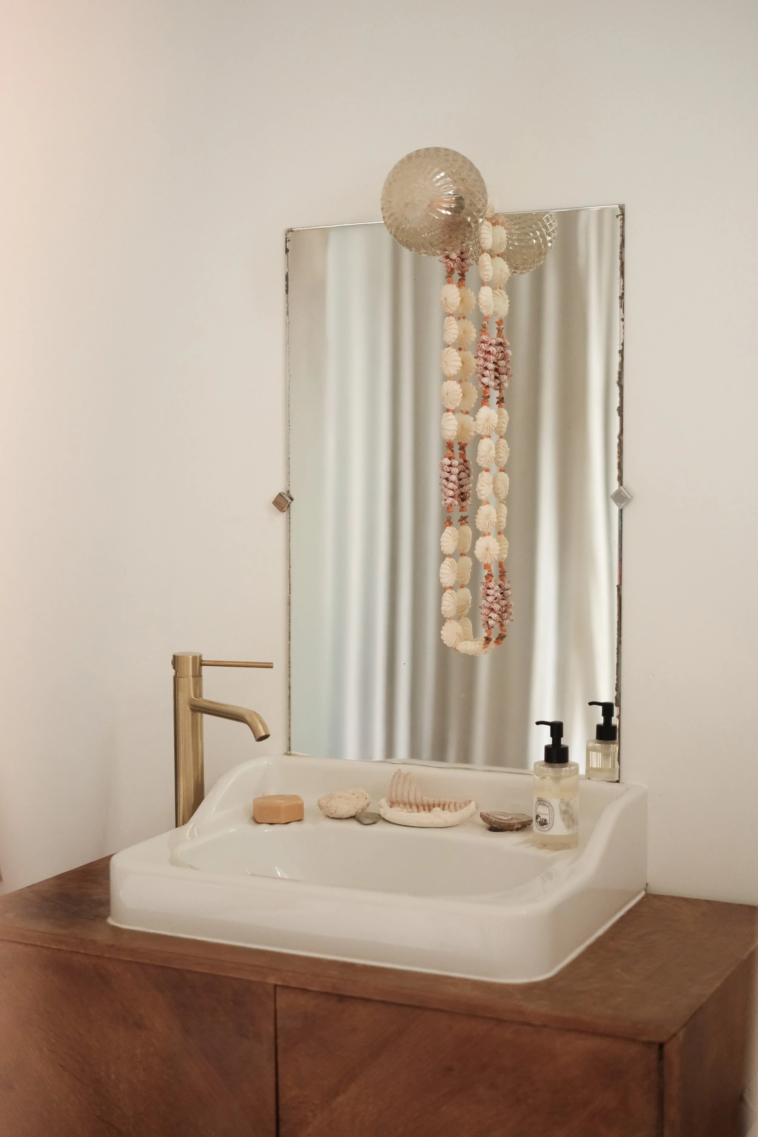 A bathroom vanity with a white sink, gold faucet, and decorative shells and stones, reflected in a mirror with hanging shell decorations and two soap dispensers.