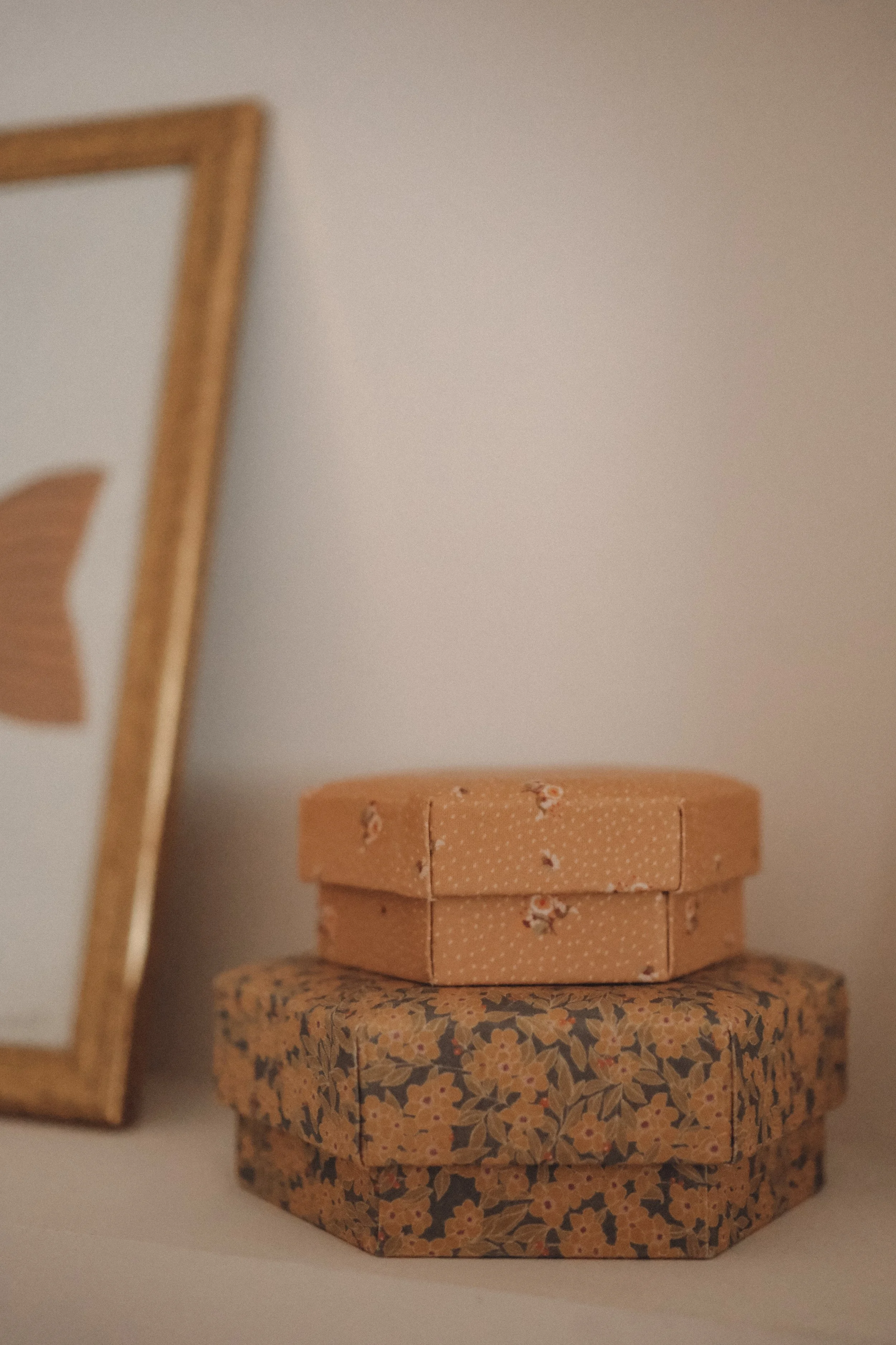 Stacked decorative boxes, one with a floral pattern and another with a dotted pattern, beside a picture frame, against a plain wall.