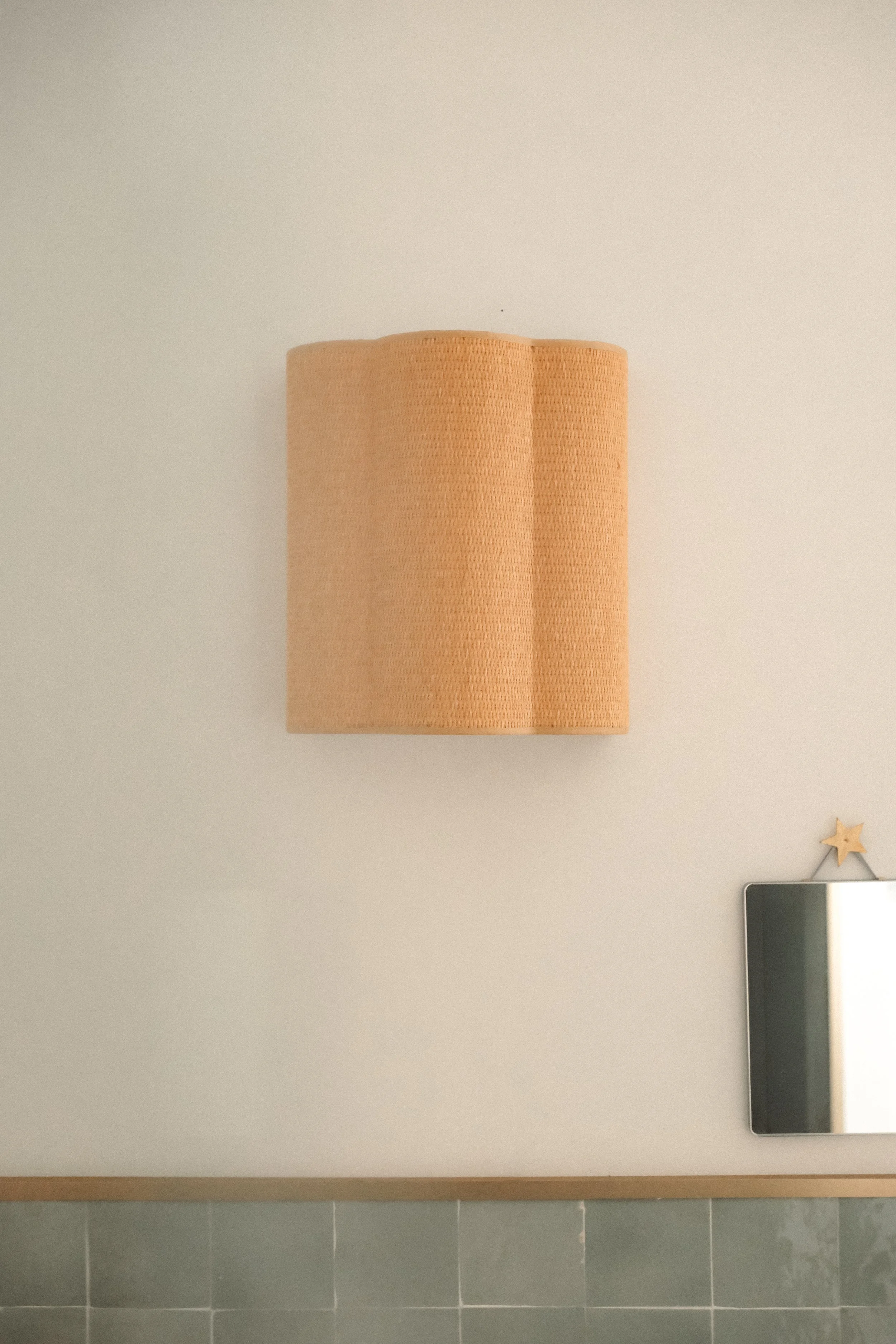 A beige wall-mounted lamp with a textured shade, positioned above a partial view of a mirror with a star-shaped decoration on top in a room with light green tiles at the bottom.
