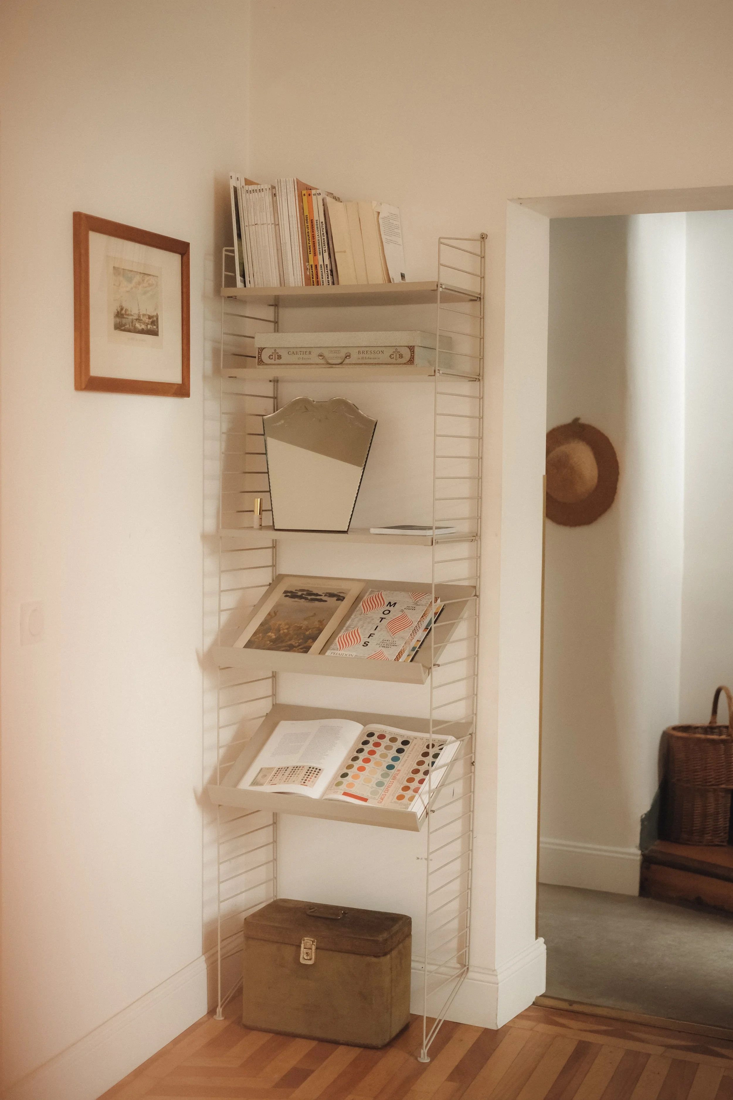 A narrow bookshelf with books and magazines, a small mirror, and decorative items against a white wall, with a small framed picture hanging beside it and a hallway with a hat on the wall nearby.