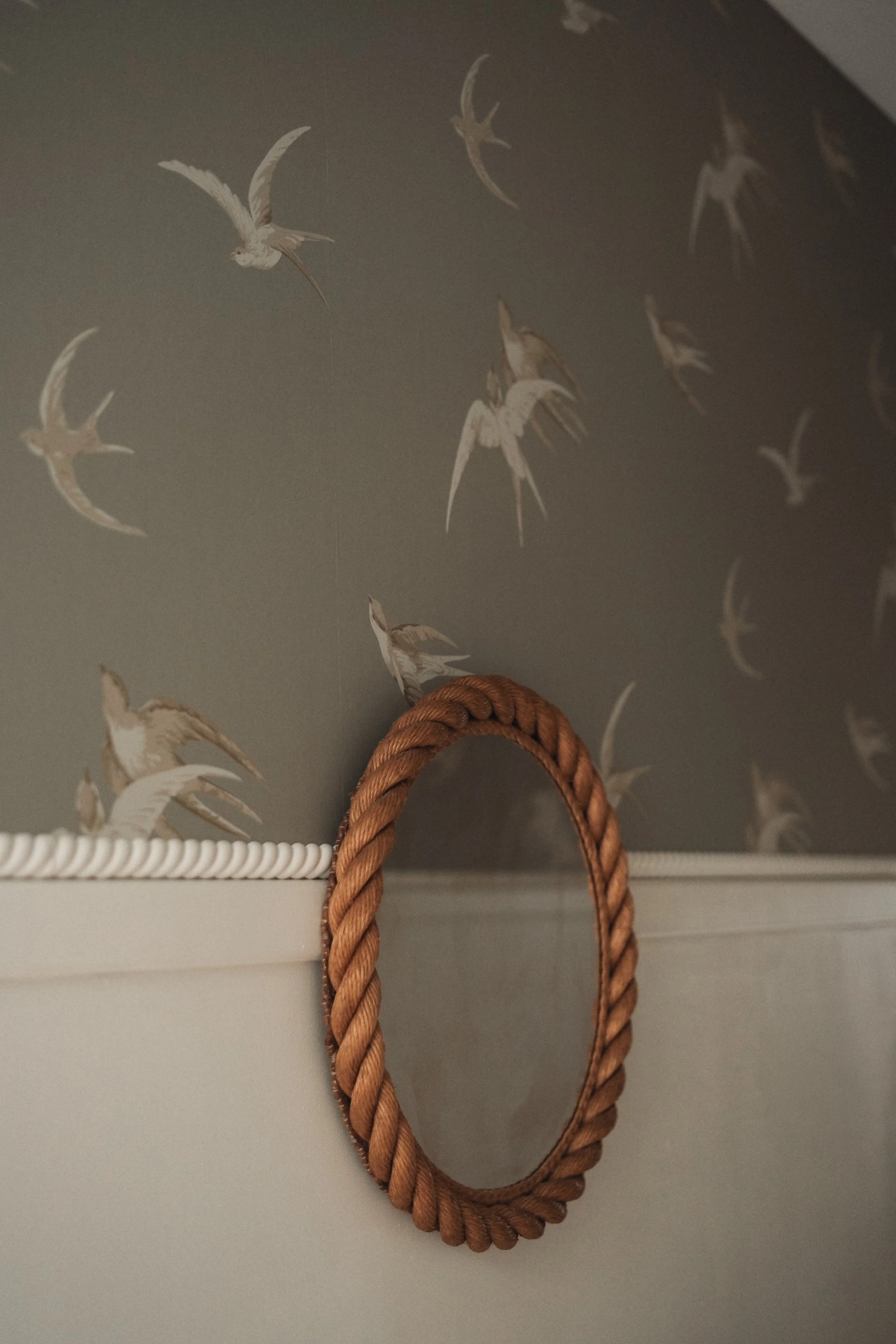 Close-up of a small oval mirror with a twisted rope frame hanging on a wall with bird-themed wallpaper.