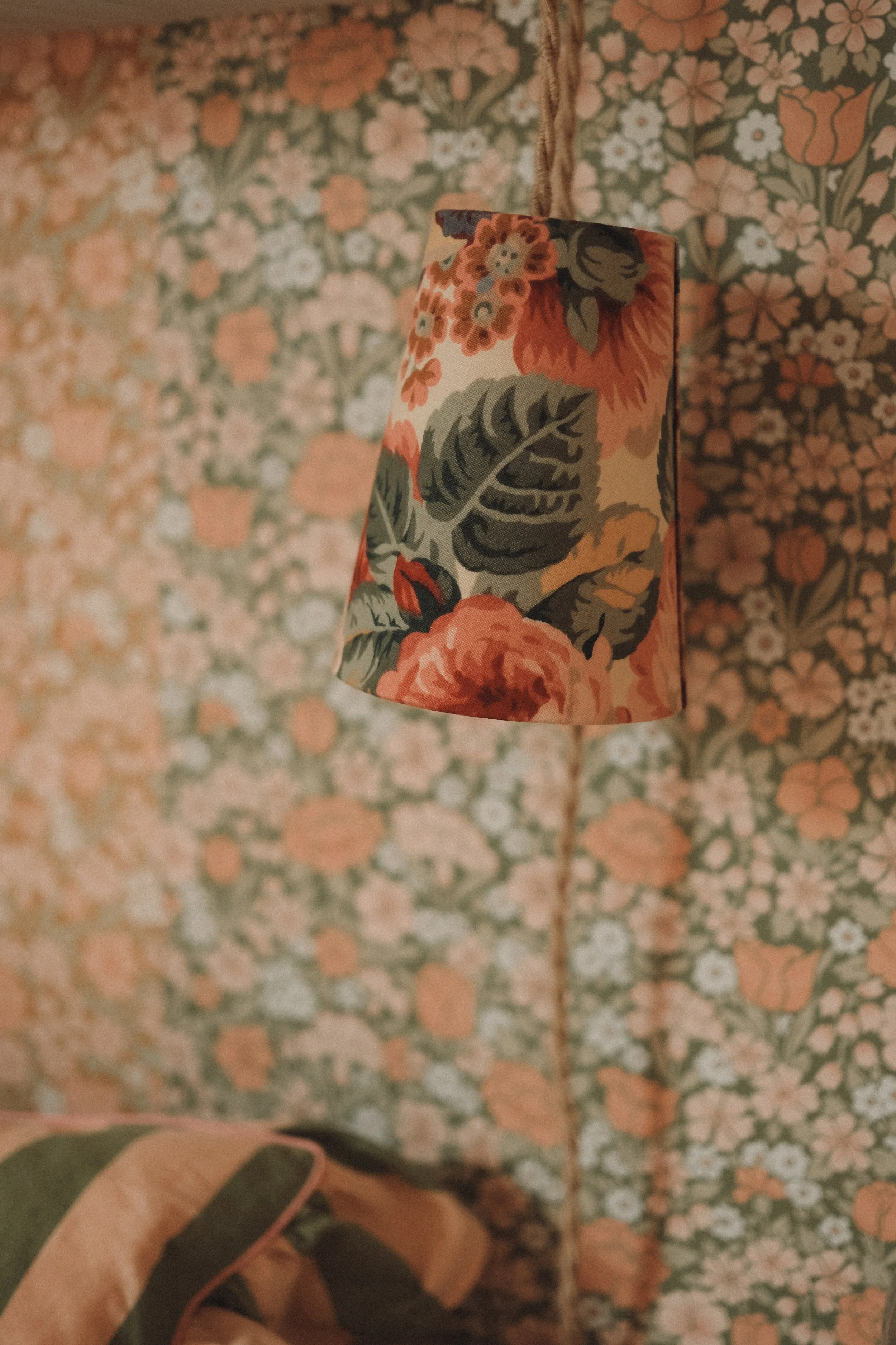 A hanging lampshade with a floral pattern of orange, red, and green leaves and flowers, against a background of similar floral wallpaper.