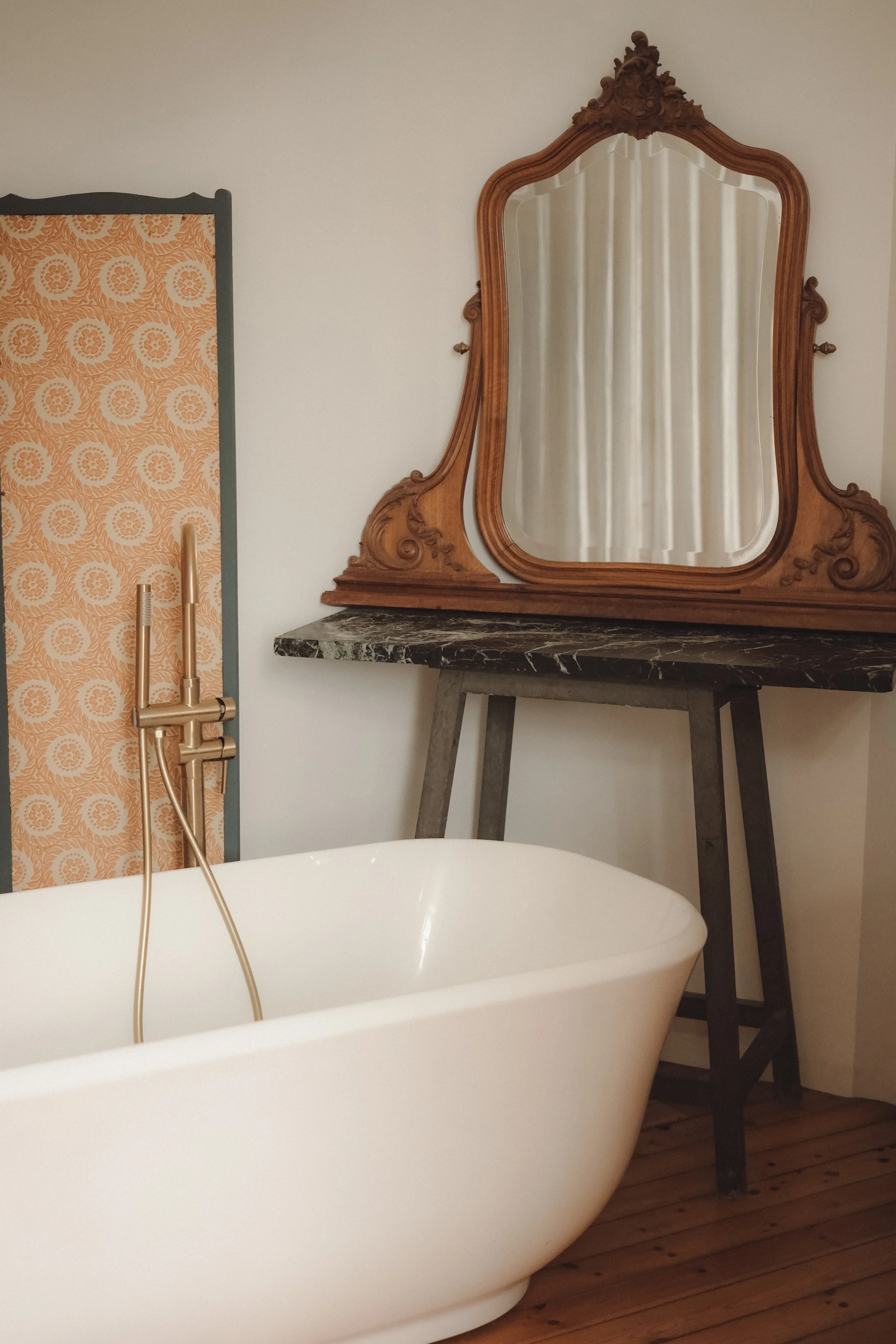 A modern white bathtub with a gold faucet is in front of an ornate wooden mirror and black marble countertop in a bathroom.