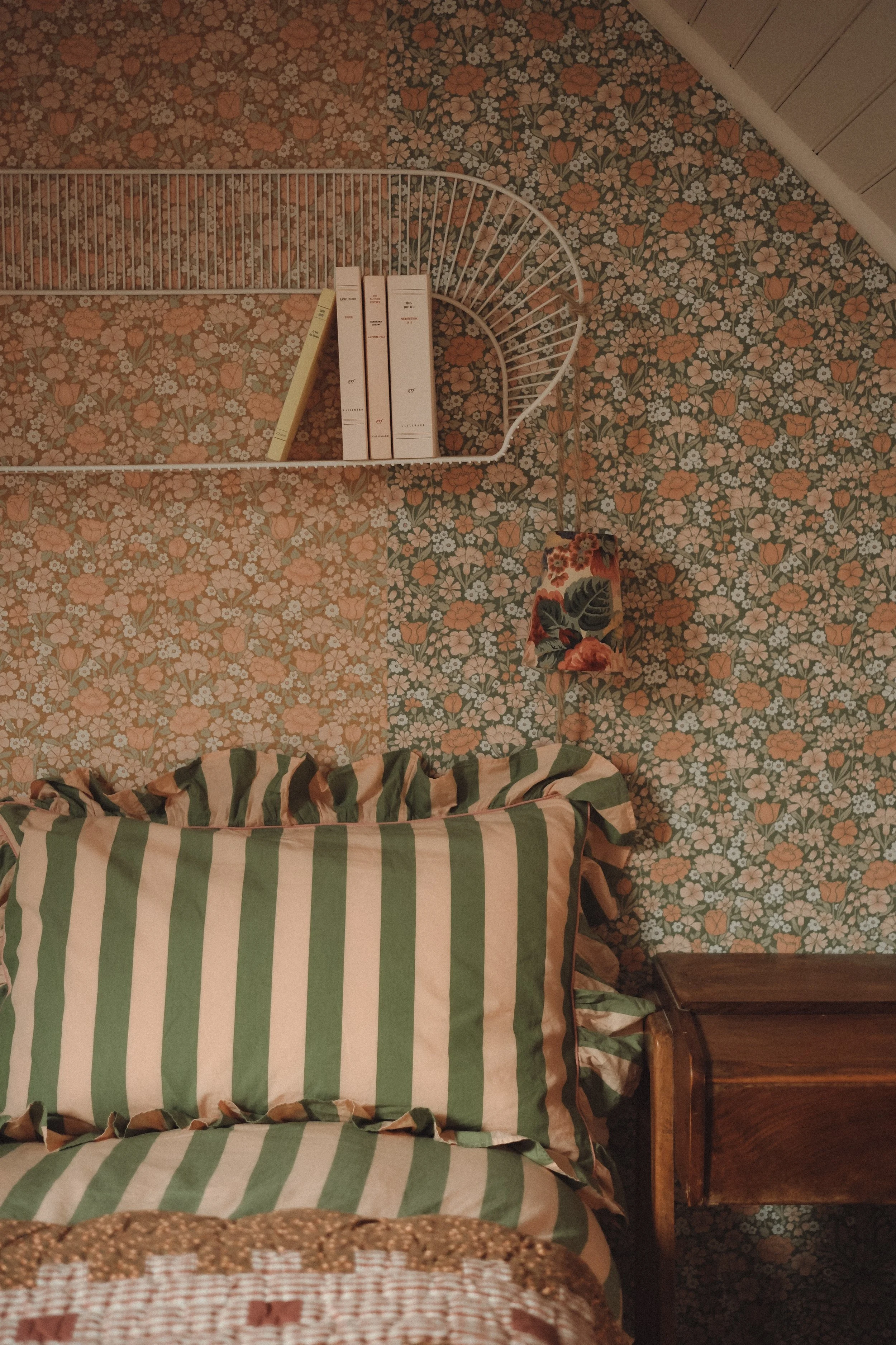A cozy bedroom with floral wallpaper, green and cream striped pillows on a bed, a small wooden nightstand, a wall-mounted wire shelf with books, and a floral lampshade.