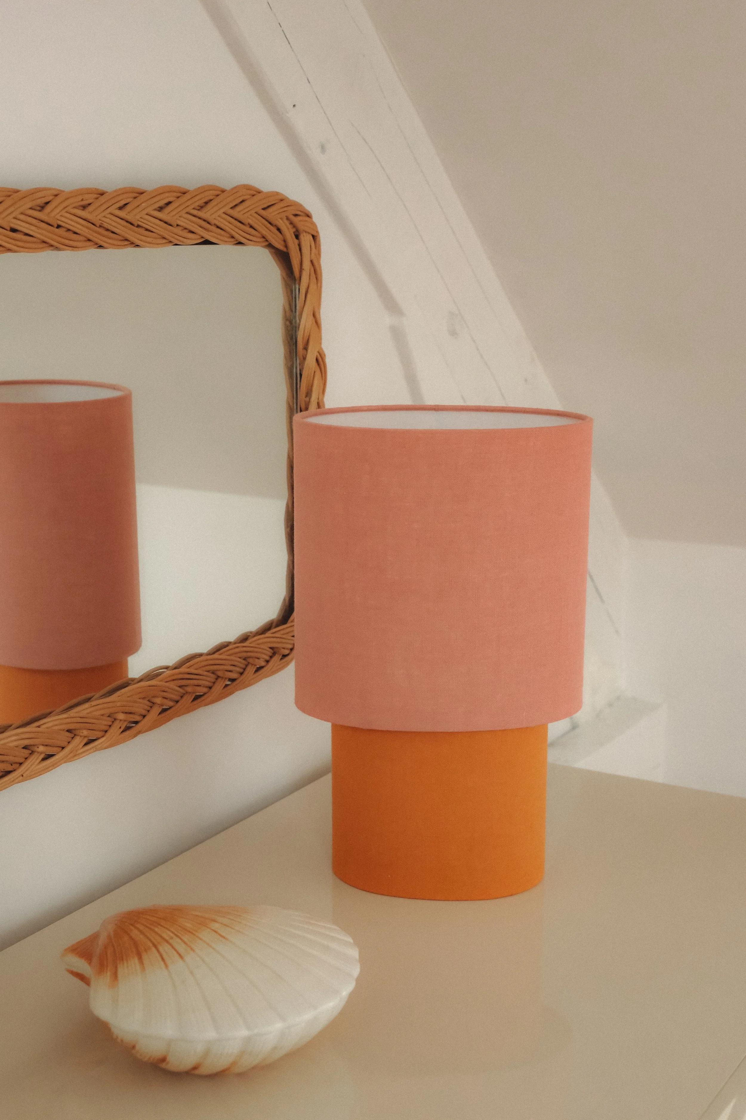 A seashell, a pink lampshade on an orange base, and a wicker-framed mirror on a white surface.