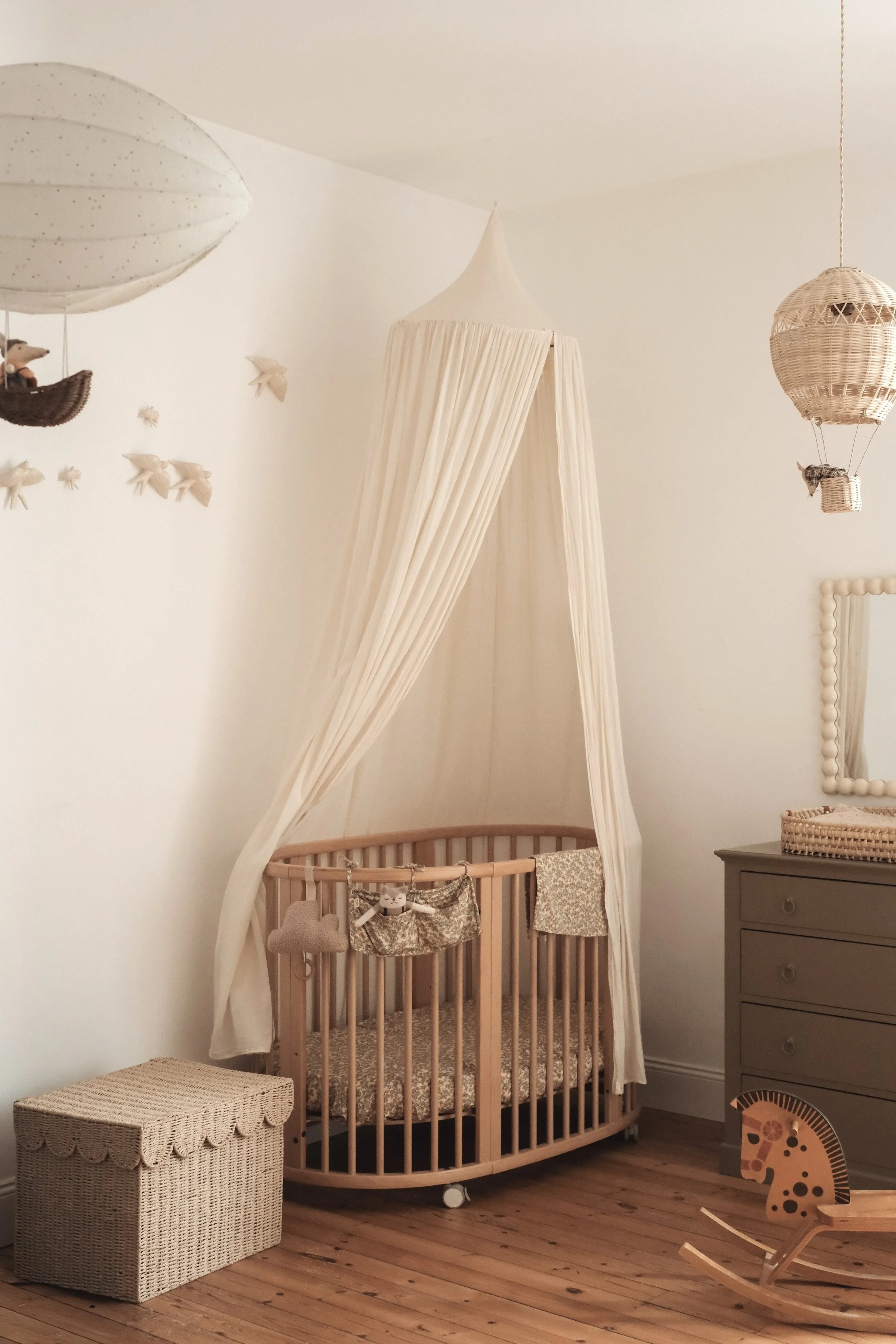 Nursery bedroom with a round wooden crib, cream canopy, hanging toys, wicker dresser with mirror, woven storage box, rocking horse, and decorated wall with small plush animals.