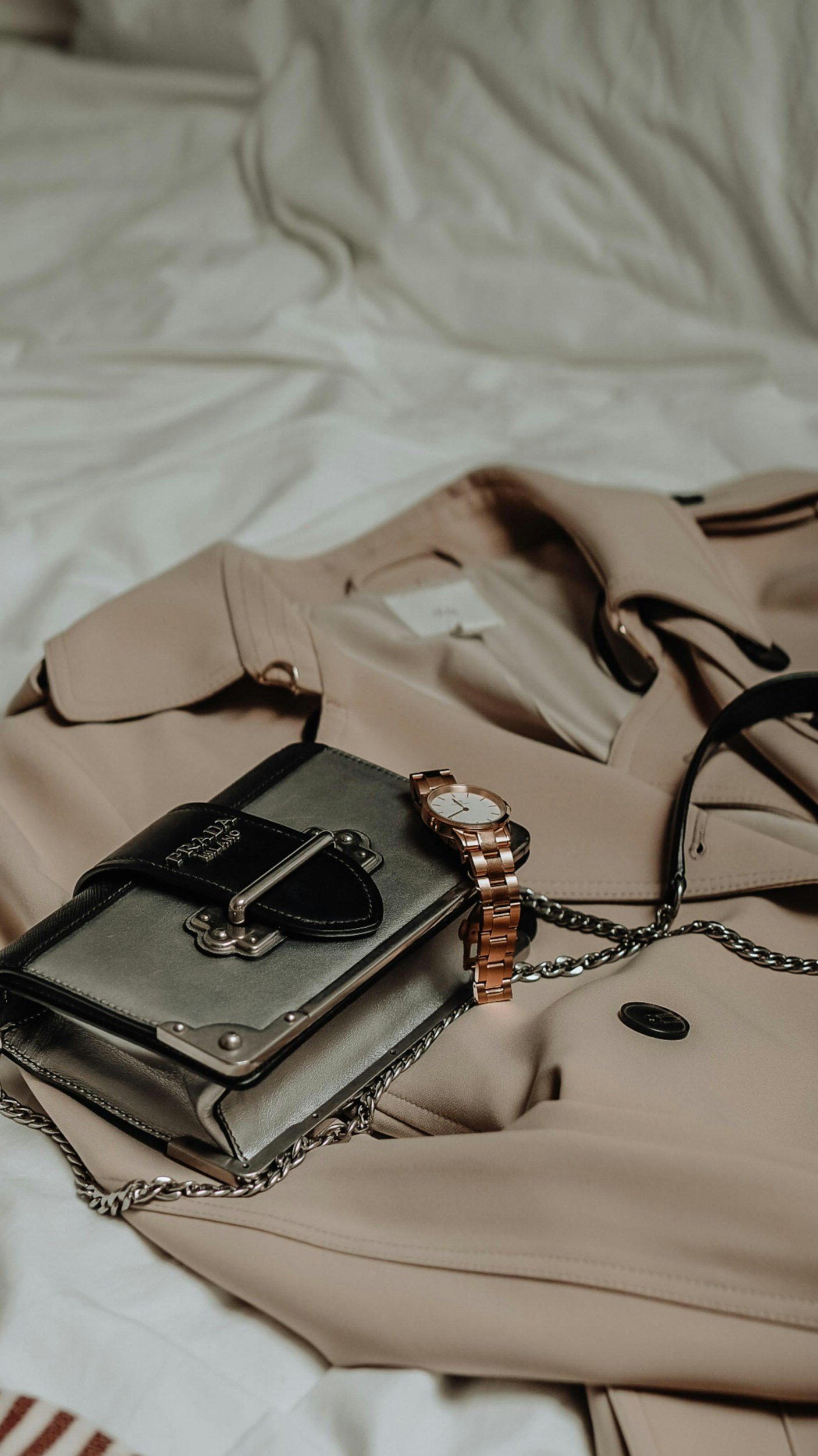 A beige trench coat laid on a bed with accessories including a metallic purse, a gold wristwatch, and a black and gold chain strap.