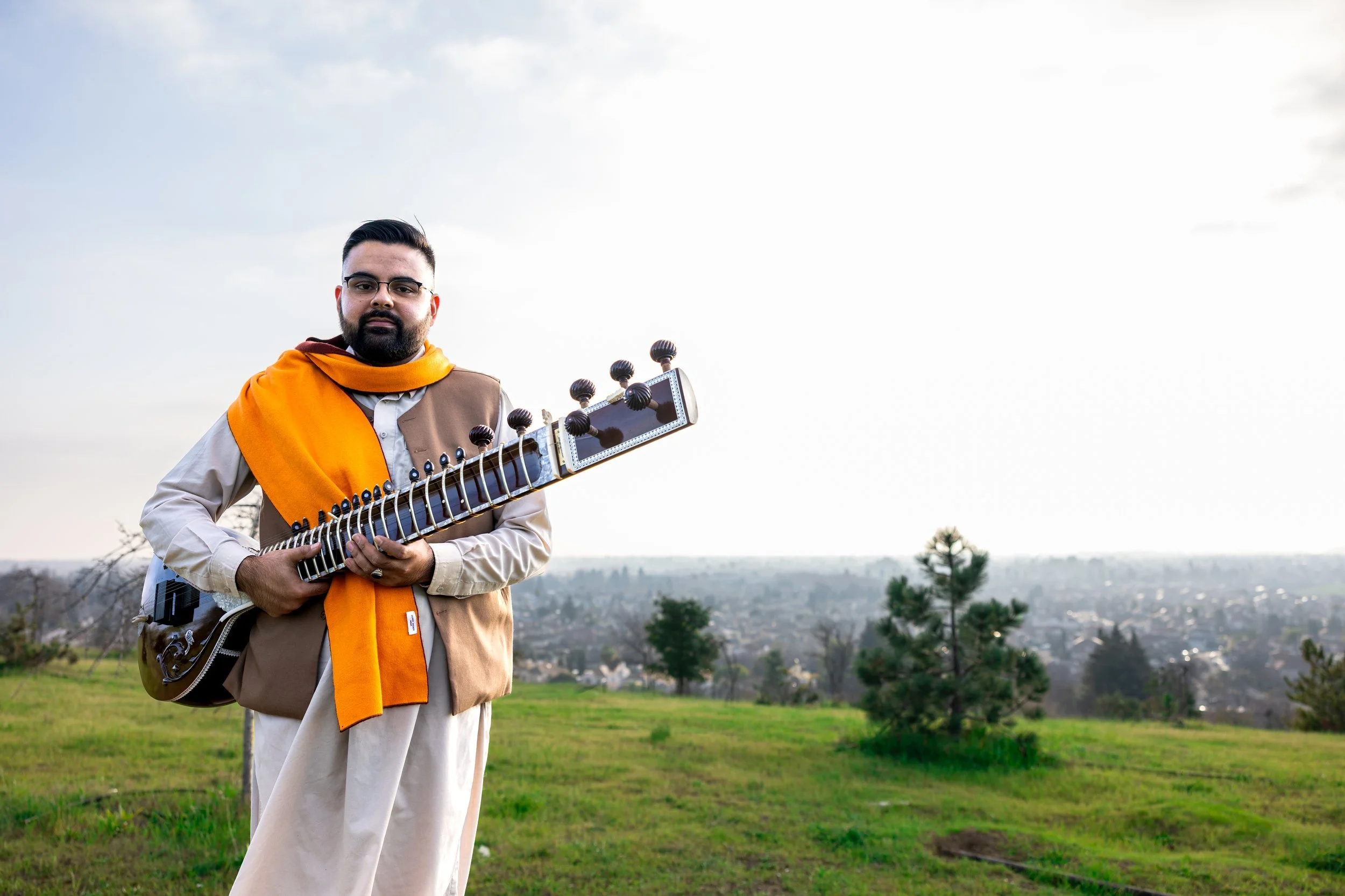 Man in traditional attire holding a sitar outdoors with a green field and cityscape in the background during daytime.