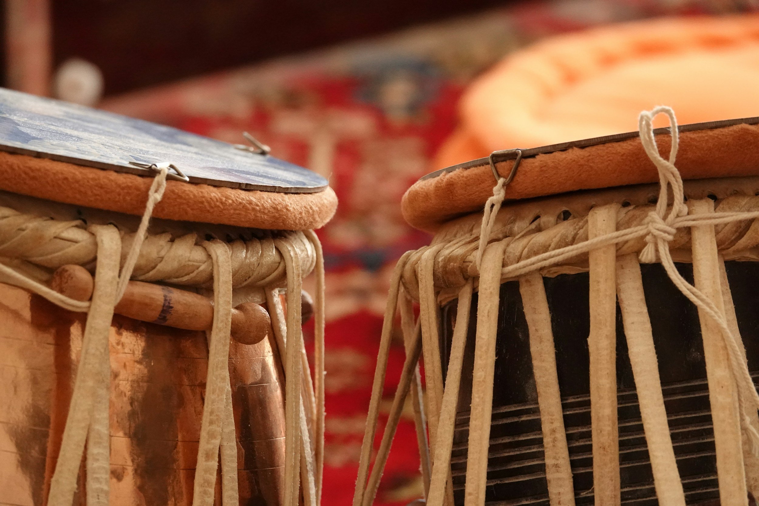 Close-up of two tabla drums with woven bodies, drumheads, and strings, set against a blurred colorful background.