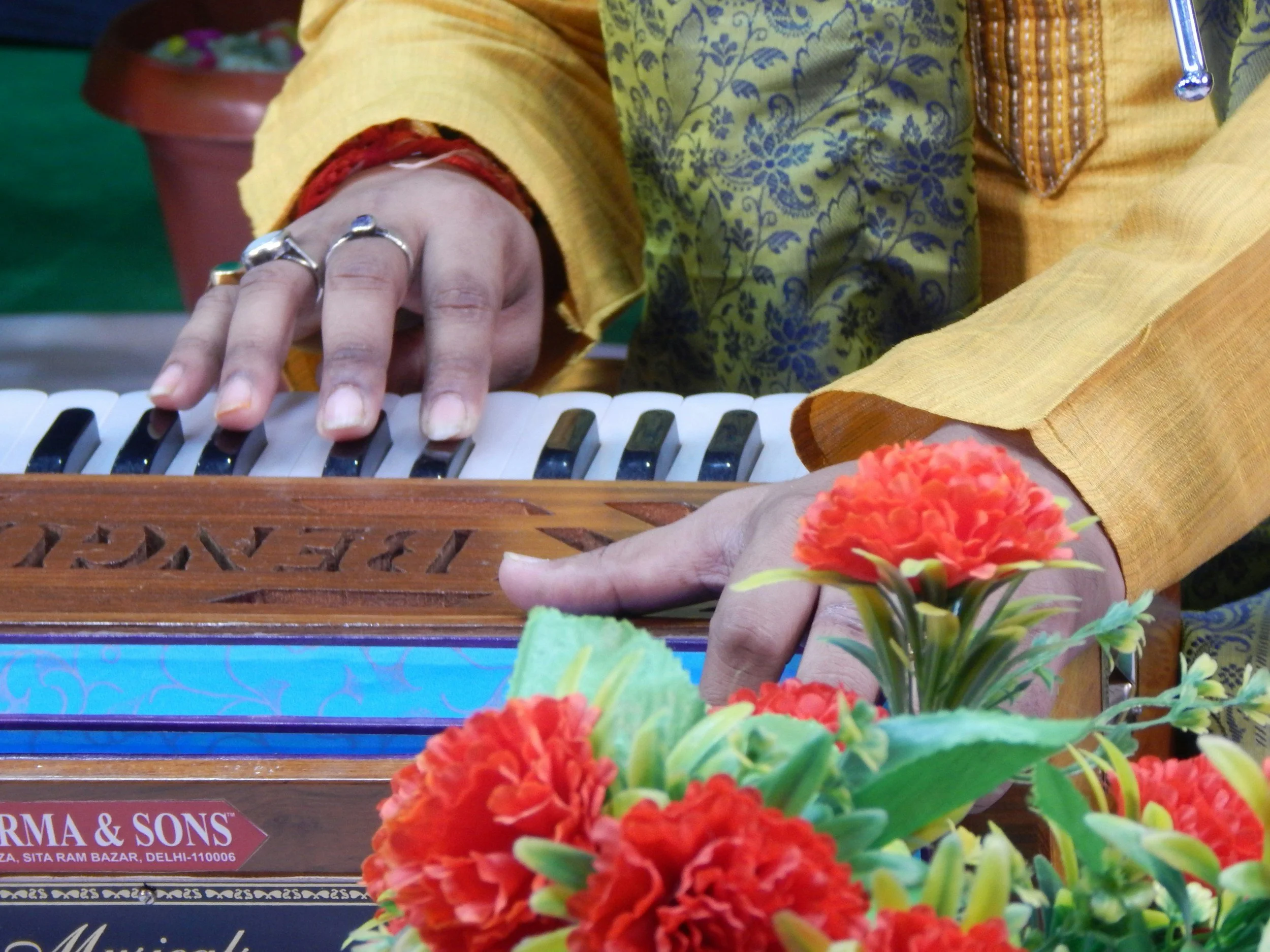 Person playing a harmonium, wearing a yellow kurta with a floral design, rings on fingers, surrounded by colorful flowers.