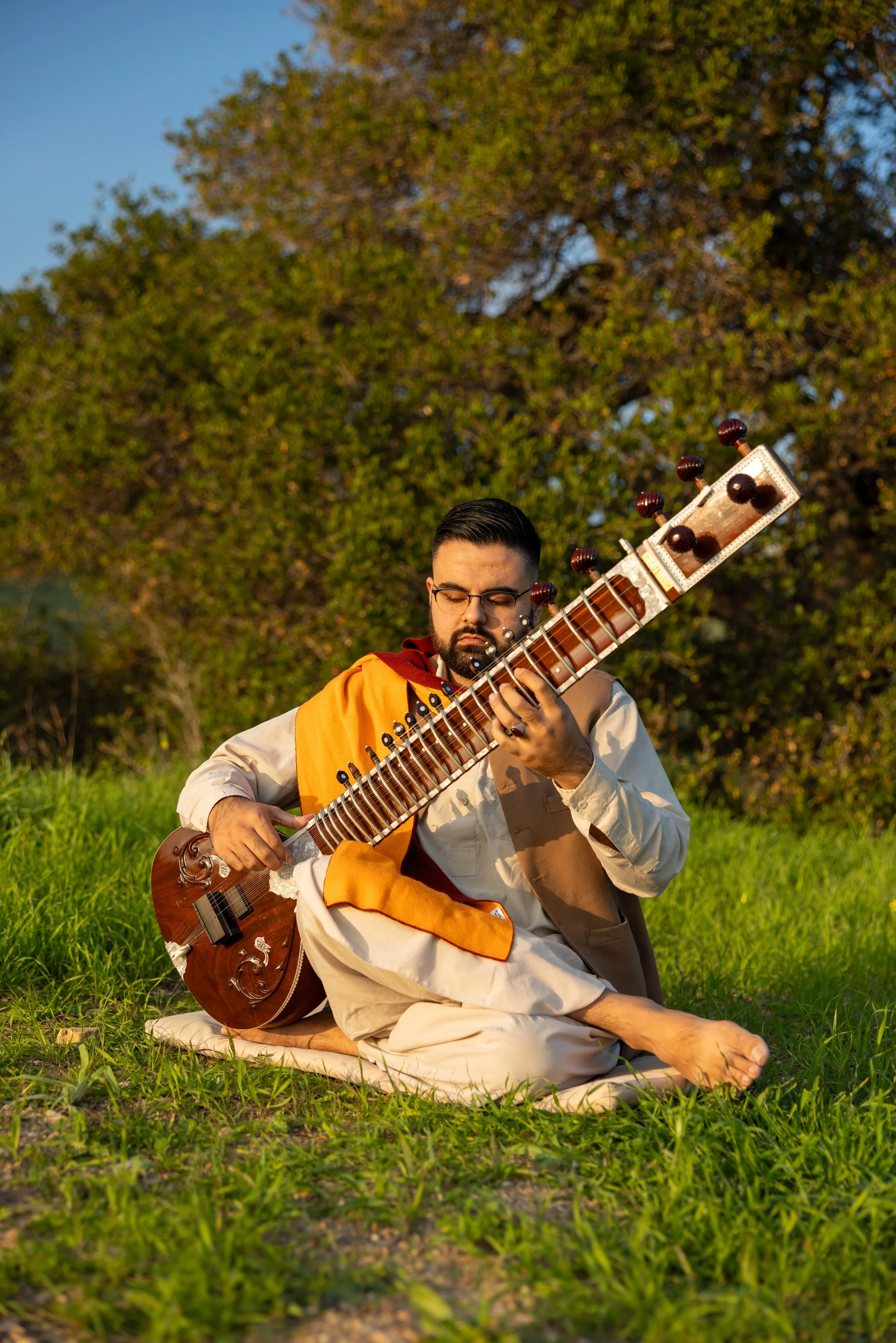 A man wearing traditional Indian attire playing a sitar outdoors in a grassy field with trees in the background during golden hour.