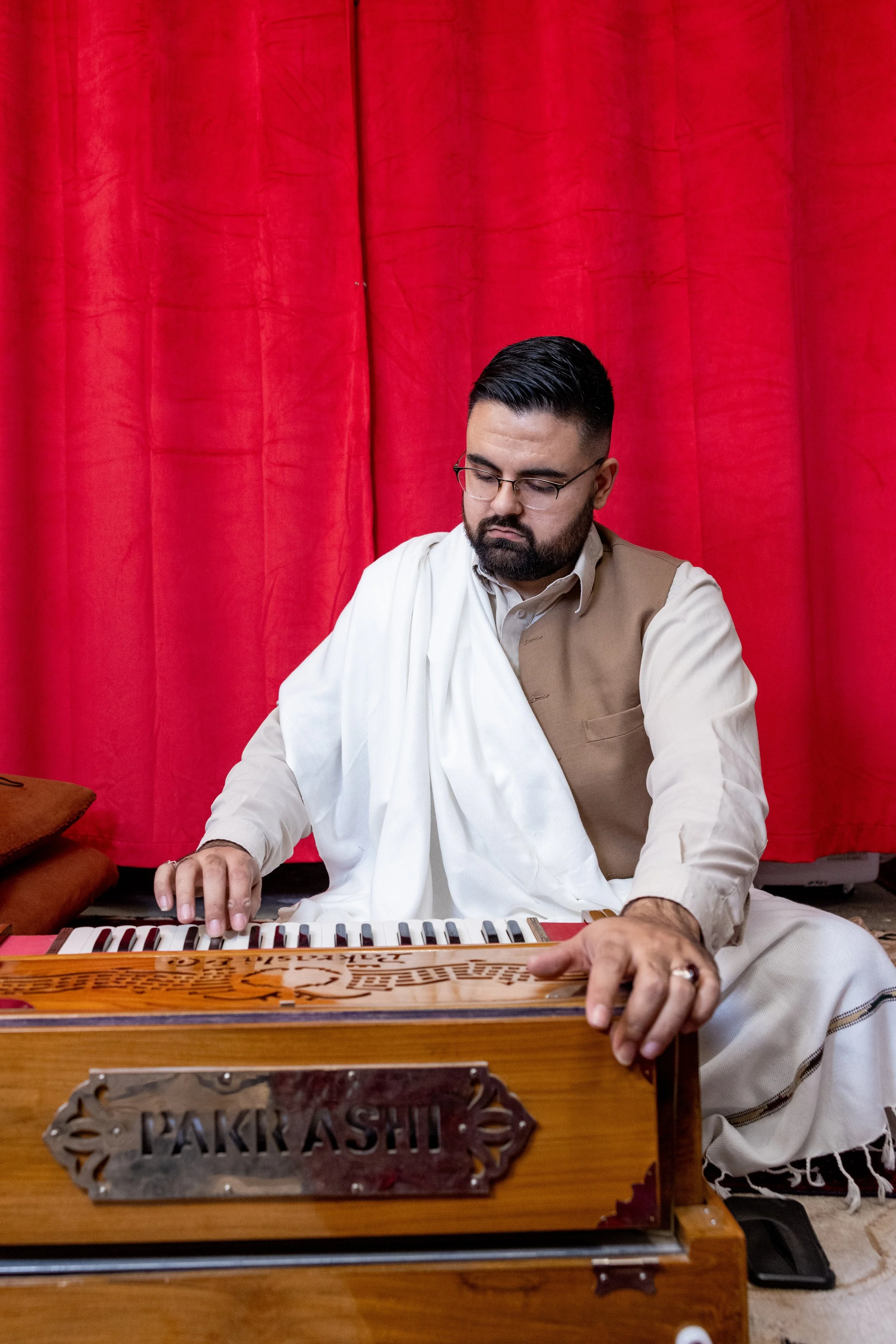 A man wearing glasses, a beige shirt, and a white traditional Indian outfit playing a harmonium in front of a red curtain.