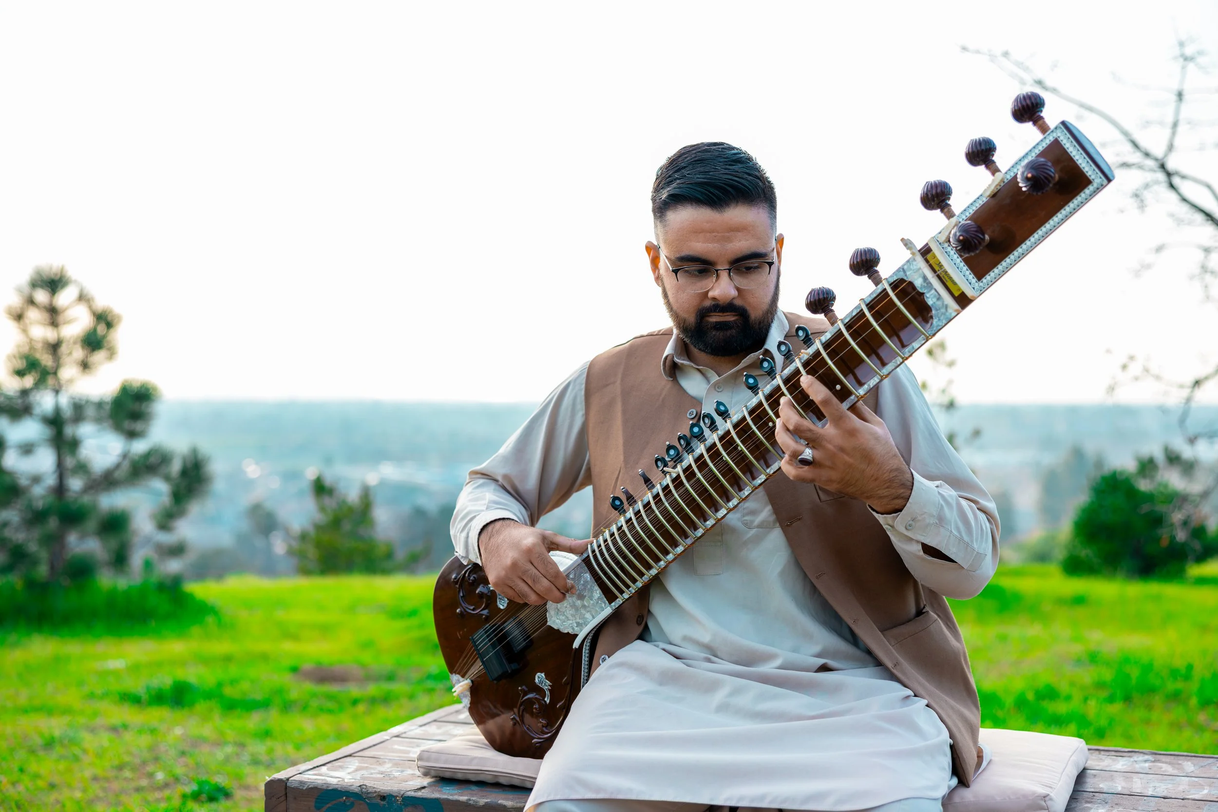 Man wearing glasses and traditional attire playing a sitar outdoors on a wooden platform with green grass and trees in the background.