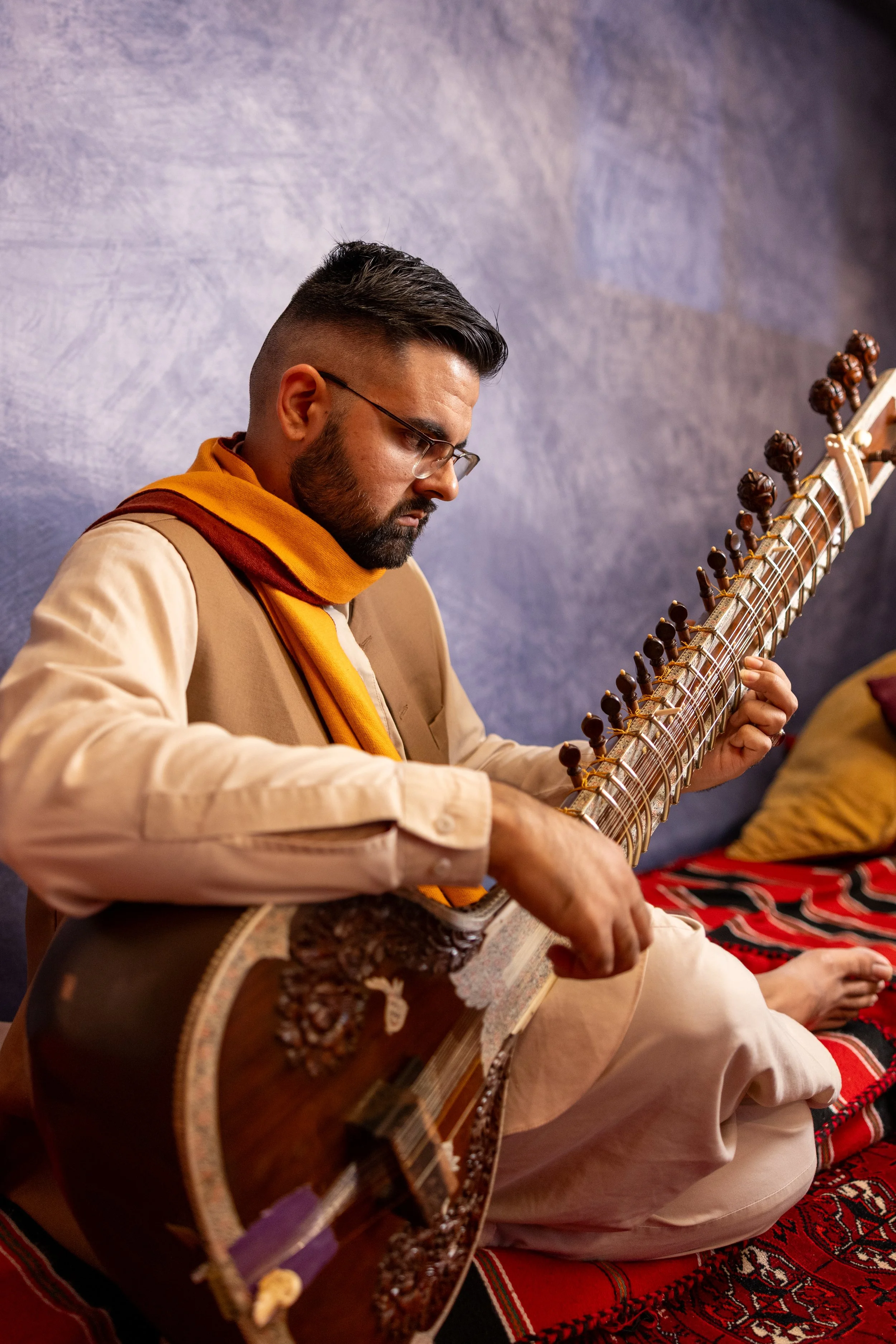 A man with glasses and a beard, wearing traditional attire including a beige kurta and a yellow-orange scarf, plays a sitar sitting on a red and black patterned rug with cushions nearby against a textured purple wall.