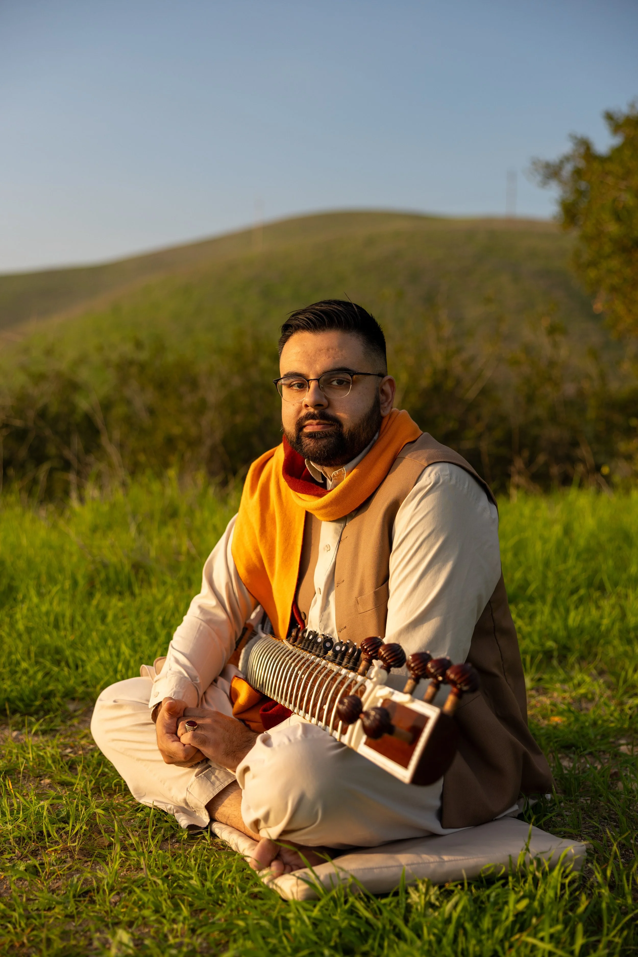 A man sitting cross-legged on grass in a natural outdoor setting during golden hour, holding a traditional musical instrument, wearing glasses, a beige traditional outfit, and a yellow and red scarf.