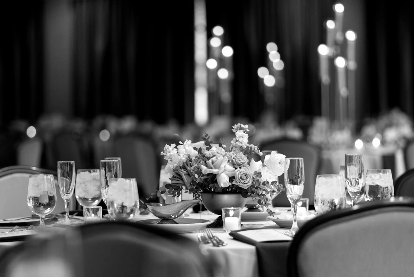 Elegant black-and-white photograph of a round table set for a formal event, with a floral centerpiece, multiple wine glasses, water glasses, and candles, surrounded by chairs, in a dimly lit room with curtains.