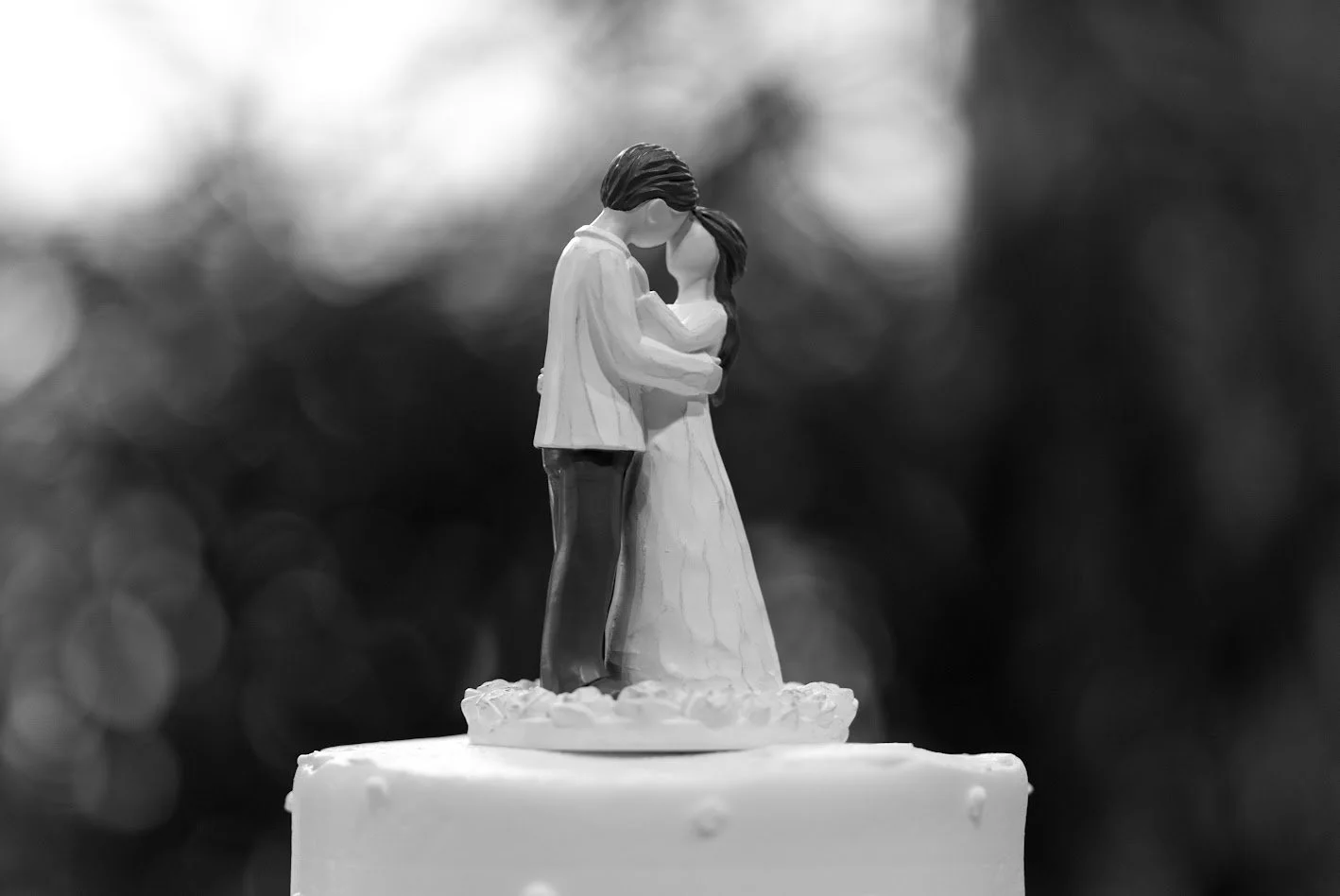 A black and white close-up of a wedding cake topped with a figurine of a bride and groom embracing.
