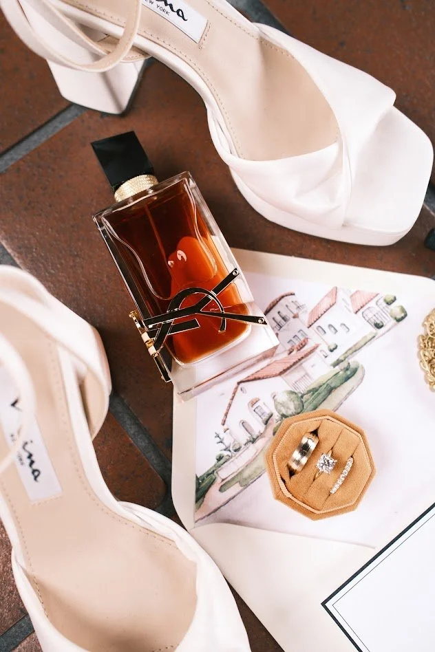 White high-heeled shoes, a bottle of perfume, a pair of sunglasses, a ring, and a diamond ring on a beige ring box, all arranged on a white cloth with a drawing of a house.