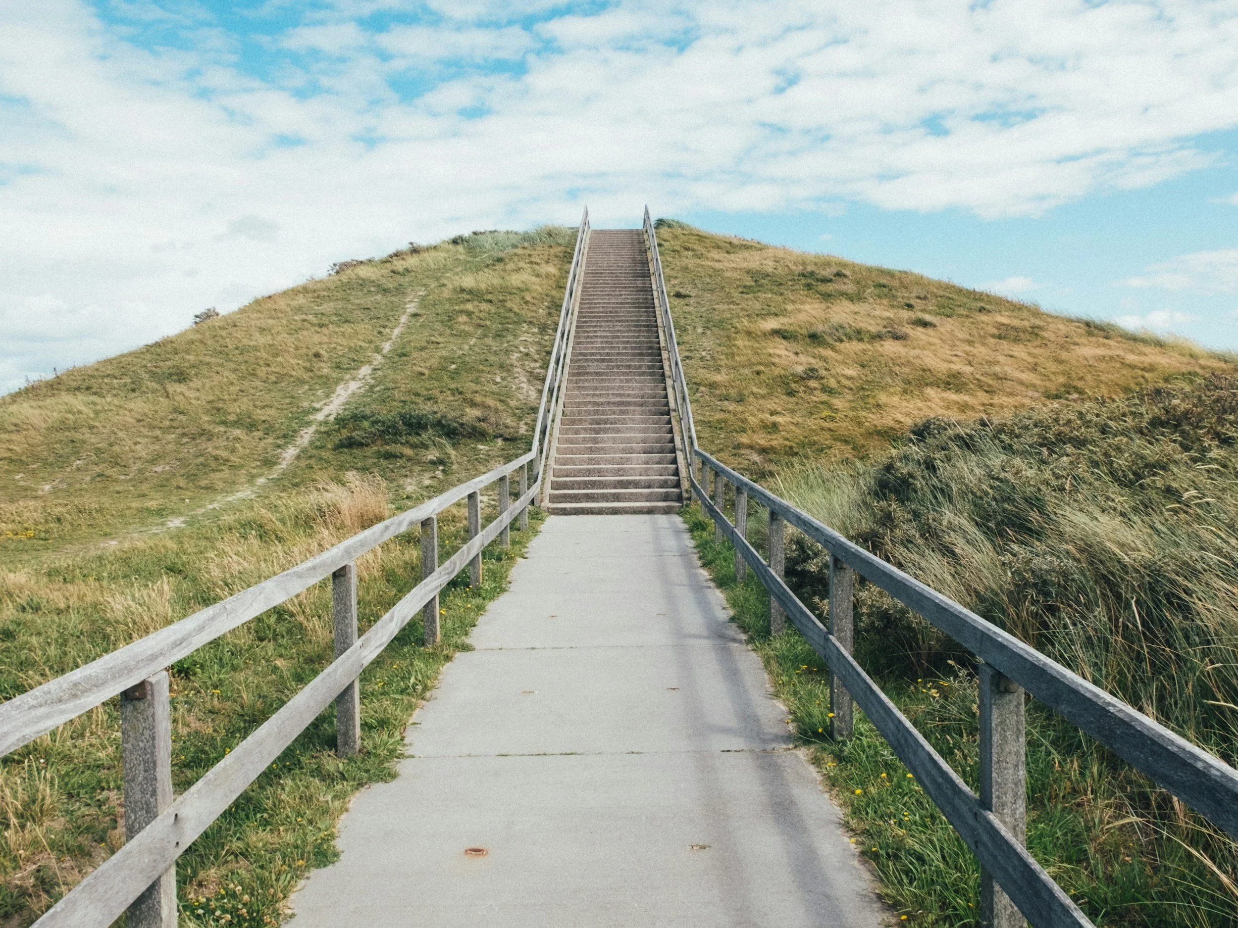 A concrete pathway leading to a staircase on a grassy hill under a blue sky with scattered white clouds.