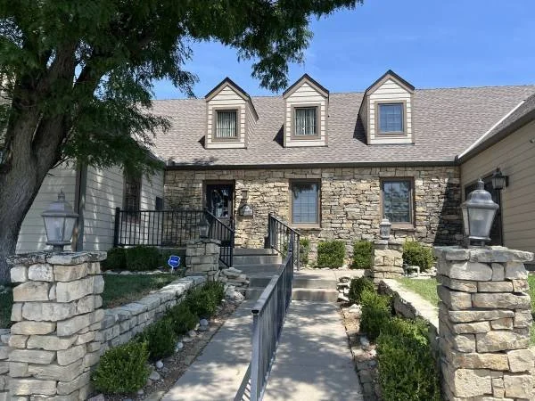 Front view of a house with a stone and siding exterior, dormer windows on the roof, and a stone pathway flanked by a stone and metal railing and landscaped bushes leading to the front door, with a large tree on the left side.
