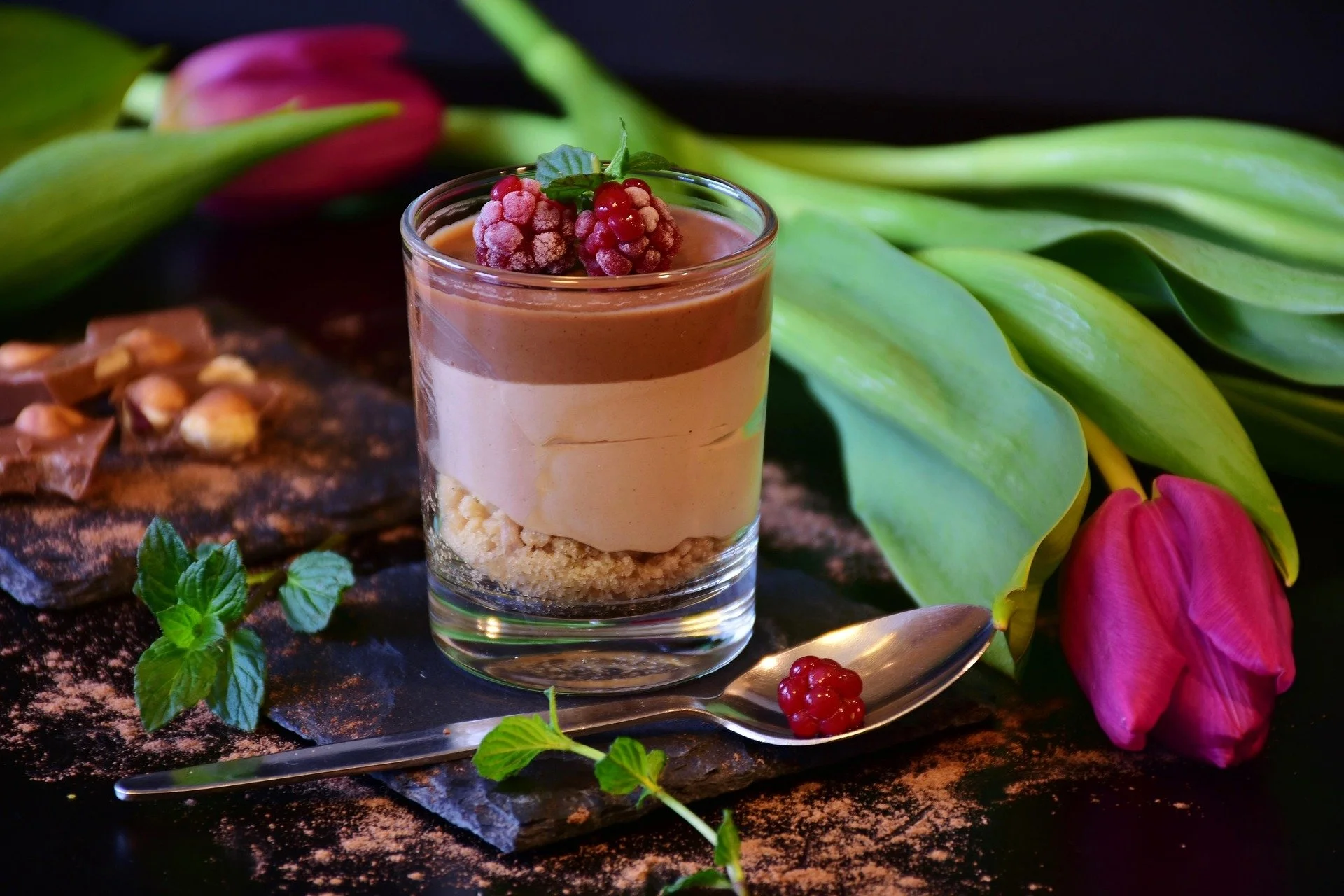 A layered dessert in a clear glass on a slate, topped with red berries and a mint leaf; surrounded by pink tulips and green leaves.