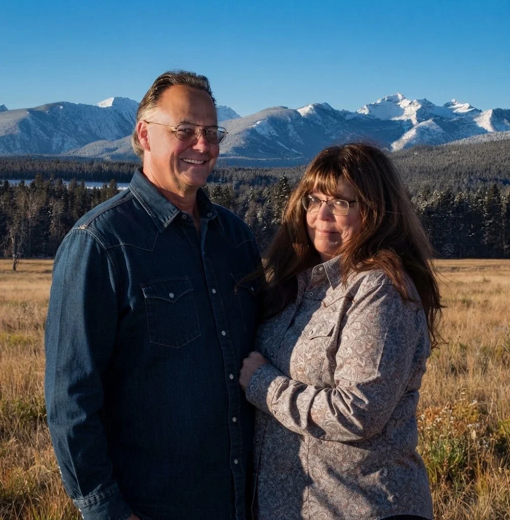 A smiling man and woman standing in a grassy field with snow-capped mountains in the background.