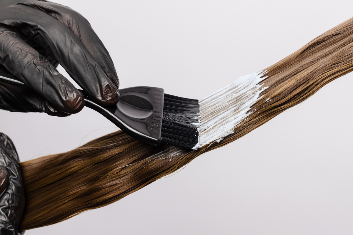 Person wearing black gloves applying hair dye to long straight brown hair with a black brush and white dye.