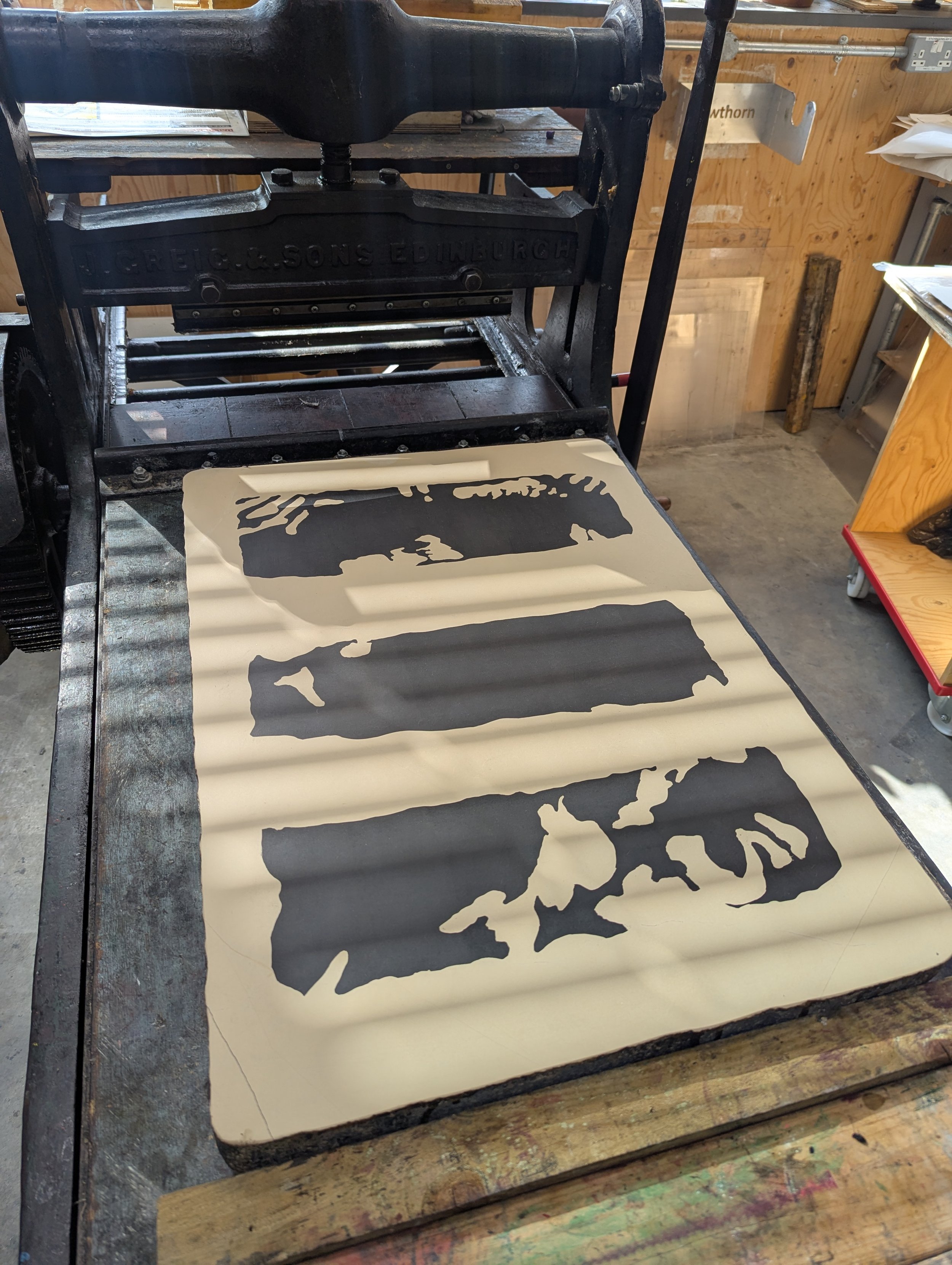 A printing press in a workshop, with a silhouette of geological forms in black on a cream-colored lithography stone ready to be printed.