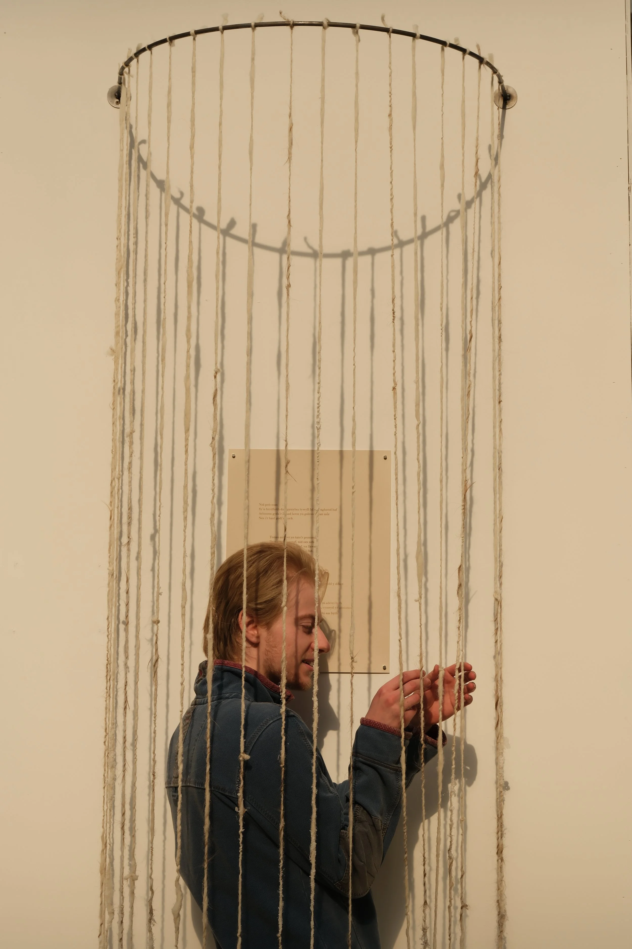 A person with light brown hair wearing a dark jacket standing behind a curtain made of strings hanging from a circular metal frame attached to the wall. The person's face is partially visible, and they appear to be adjusting or holding the strings.