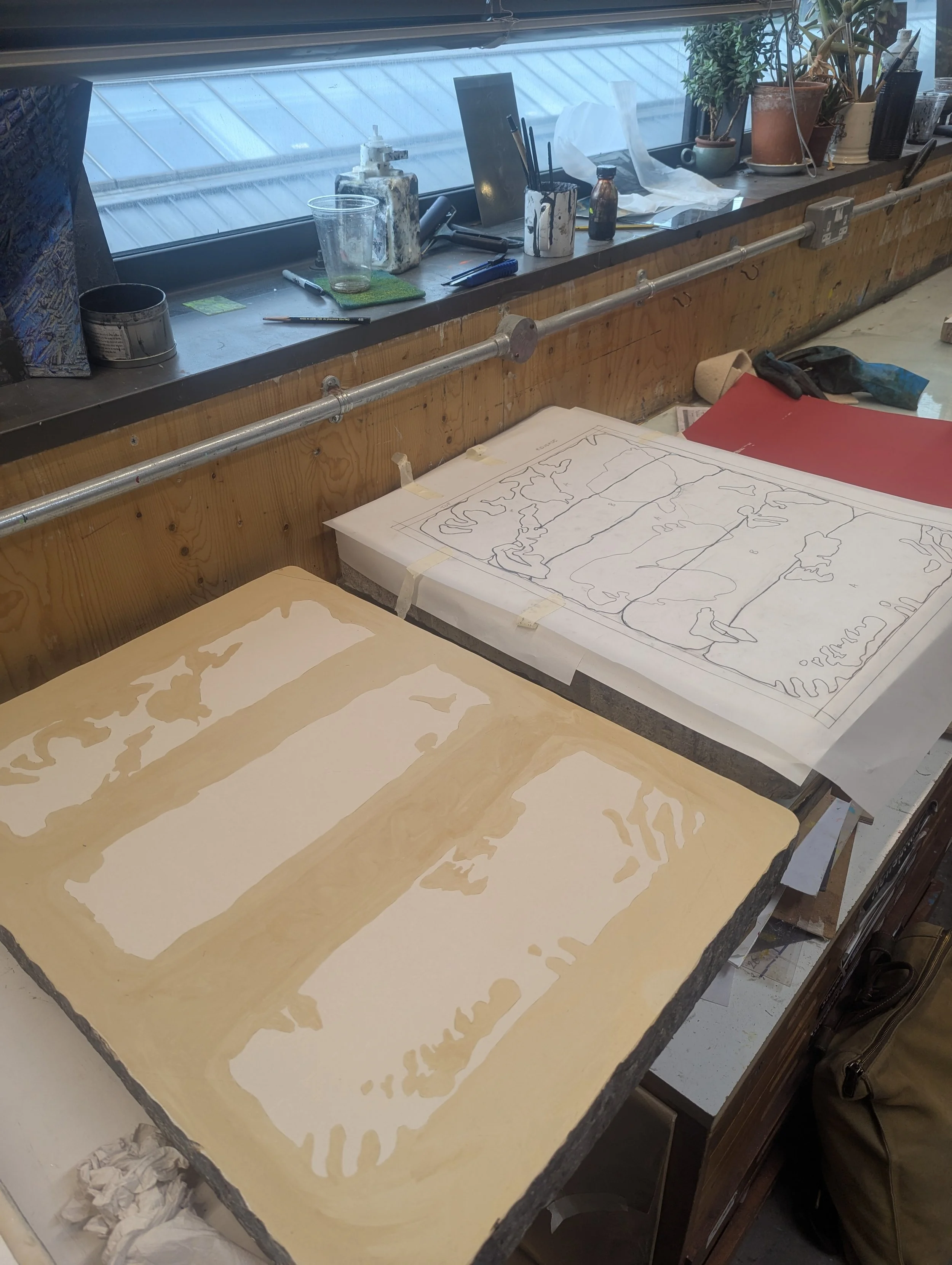 Print workshop with large lithography stones on a workbench and pencil sketches, beside a windowsill cluttered with art supplies, pens, potted plants, and containers, near a large window.