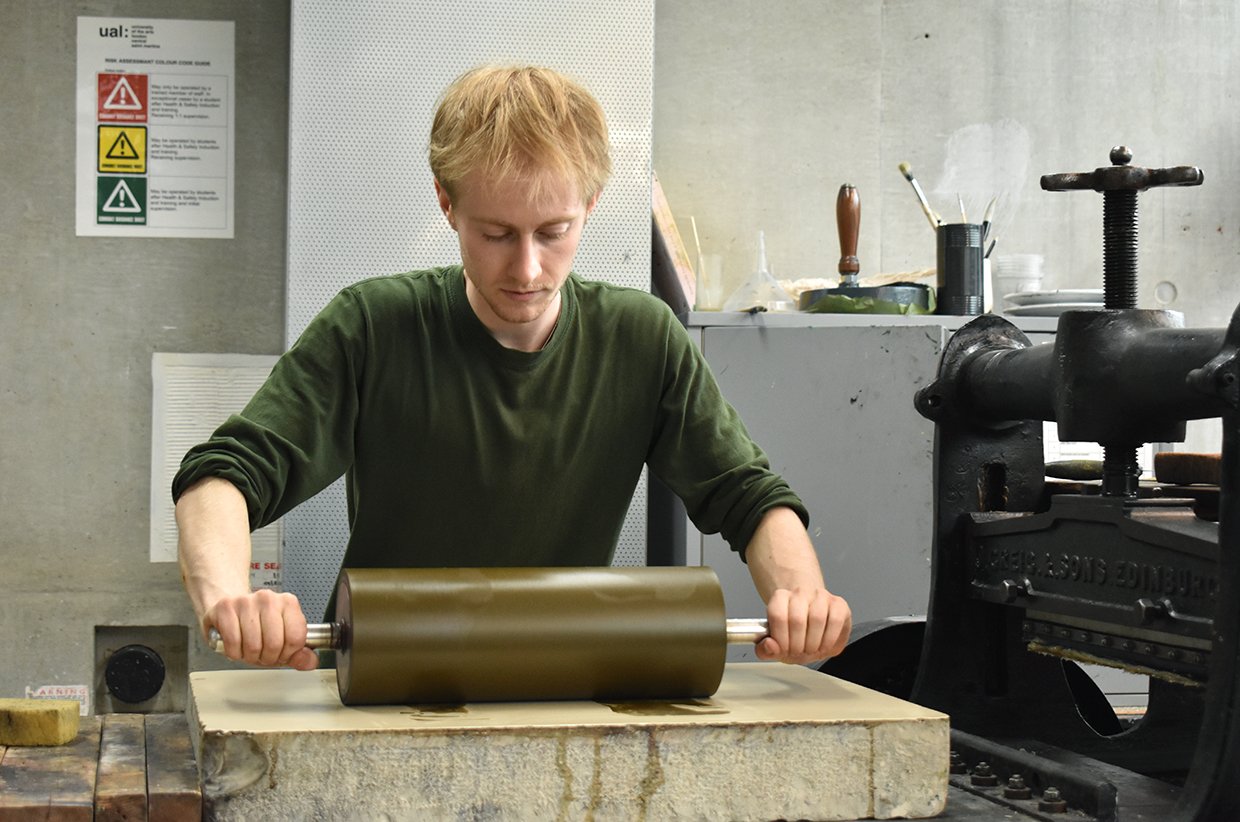 A man in a green shirt using a roller with a handle on a large stone slab in a workshop setting.