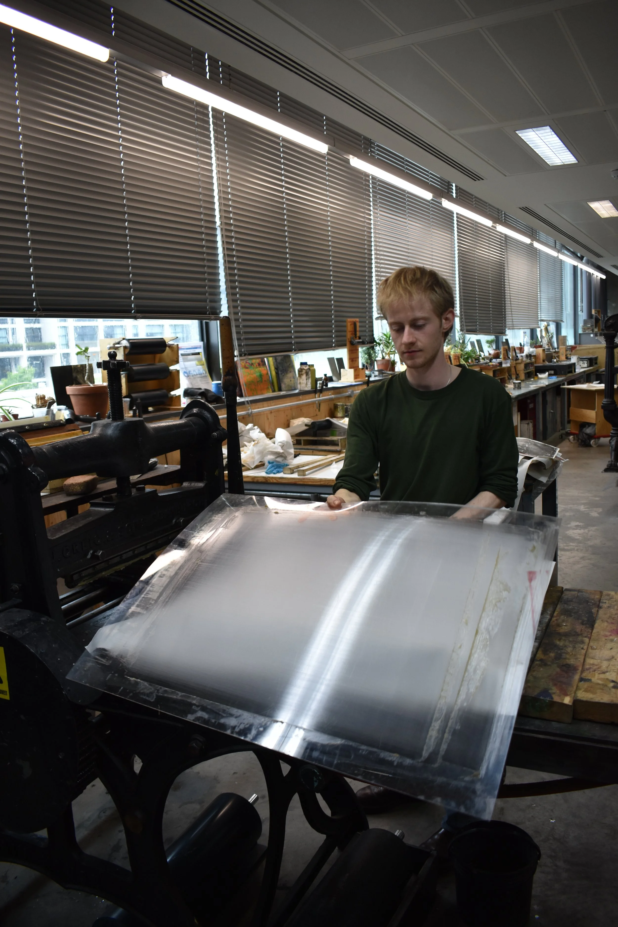 A young man in a green shirt working in a workshop or studio, handling a large sheet of shiny, reflective material, with various tools and supplies on the workbench nearby, and large windows with blinds in the background.