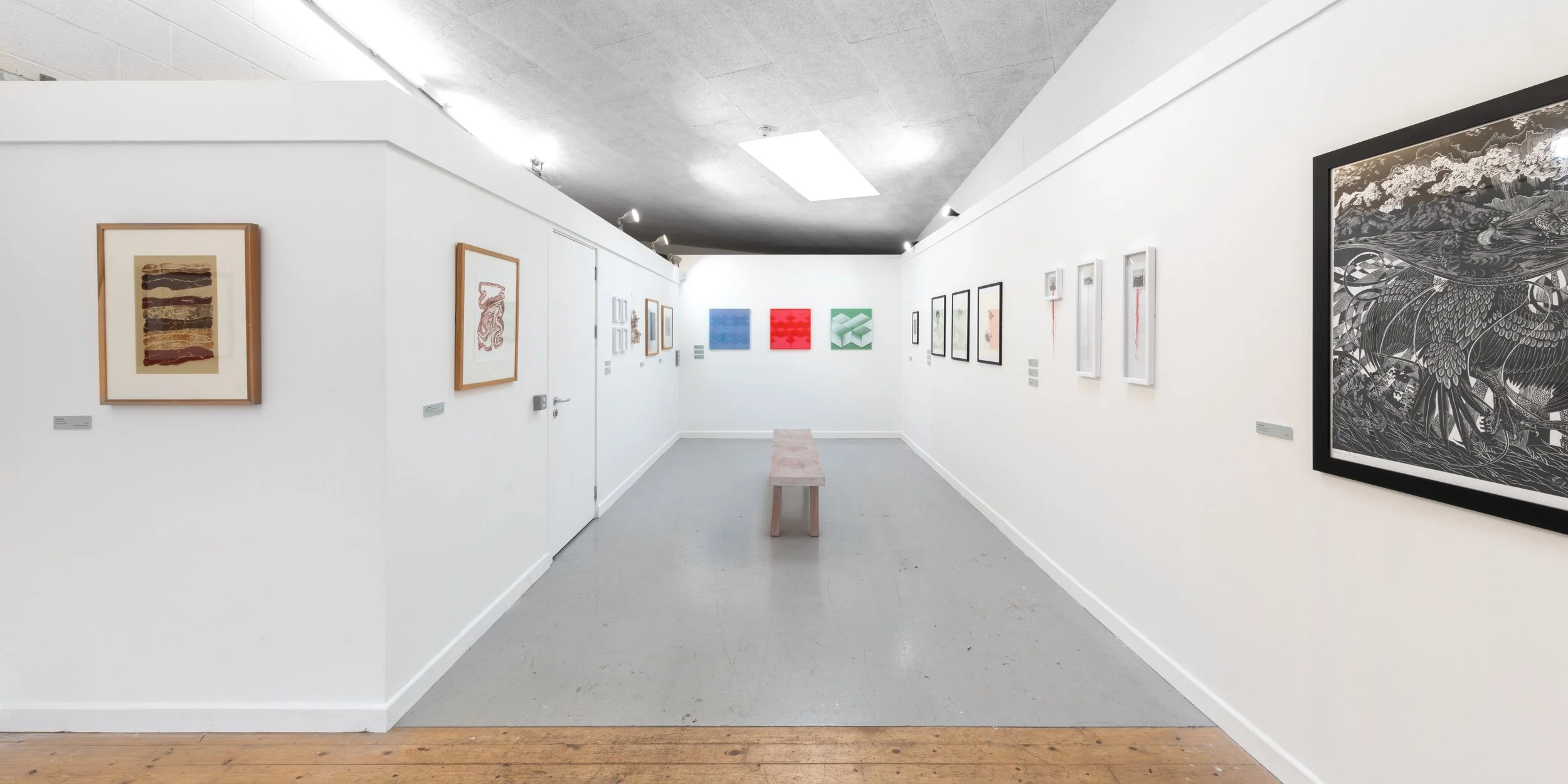 A modern art gallery with white walls displaying various framed artworks, a pink bench in the center, and a ceiling with fluorescent lighting.