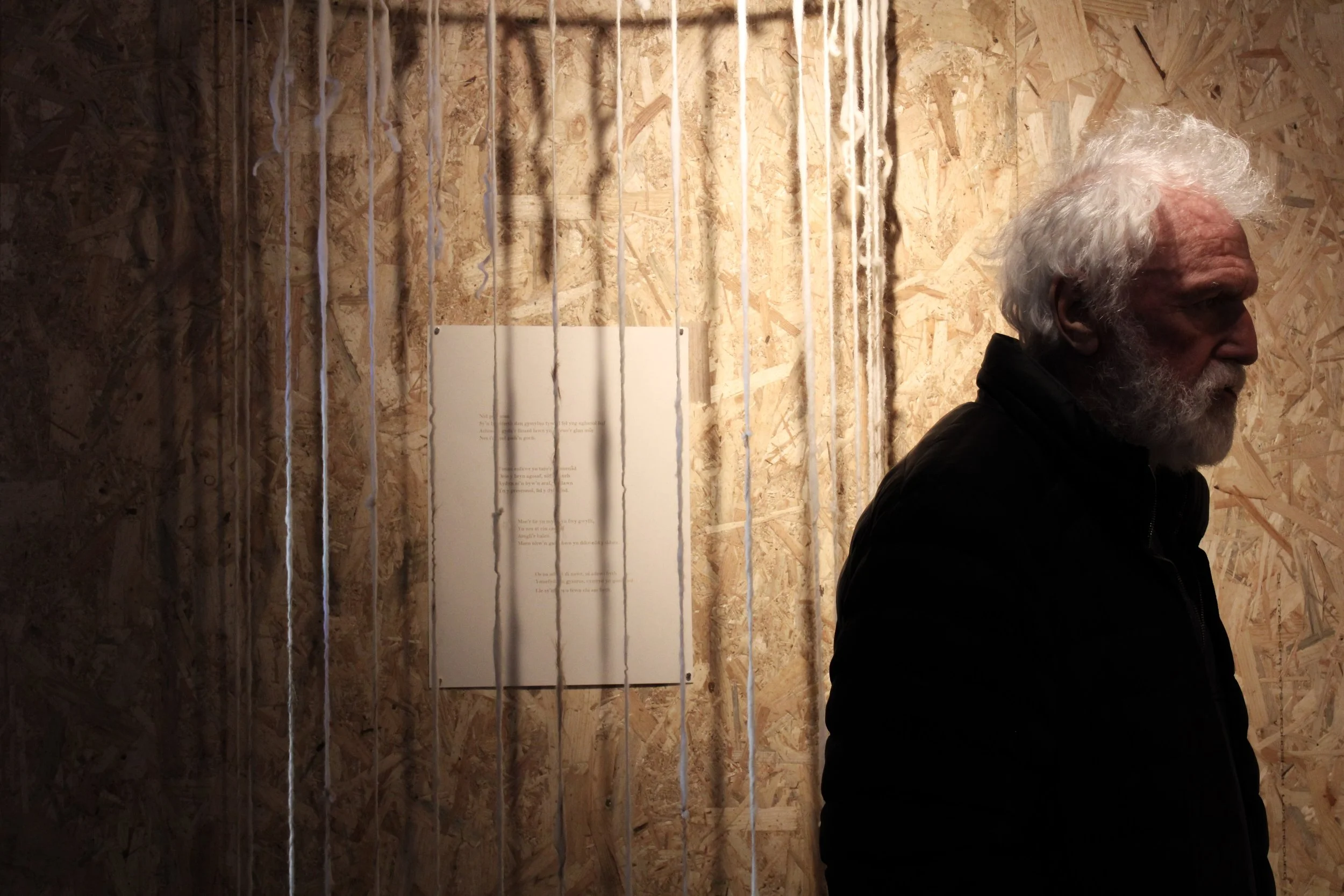 A man with white, curly hair and a beard standing in front of a wooden wall with poetic text and hanging strings or wires, partially obscured by shadows and a dark jacket.