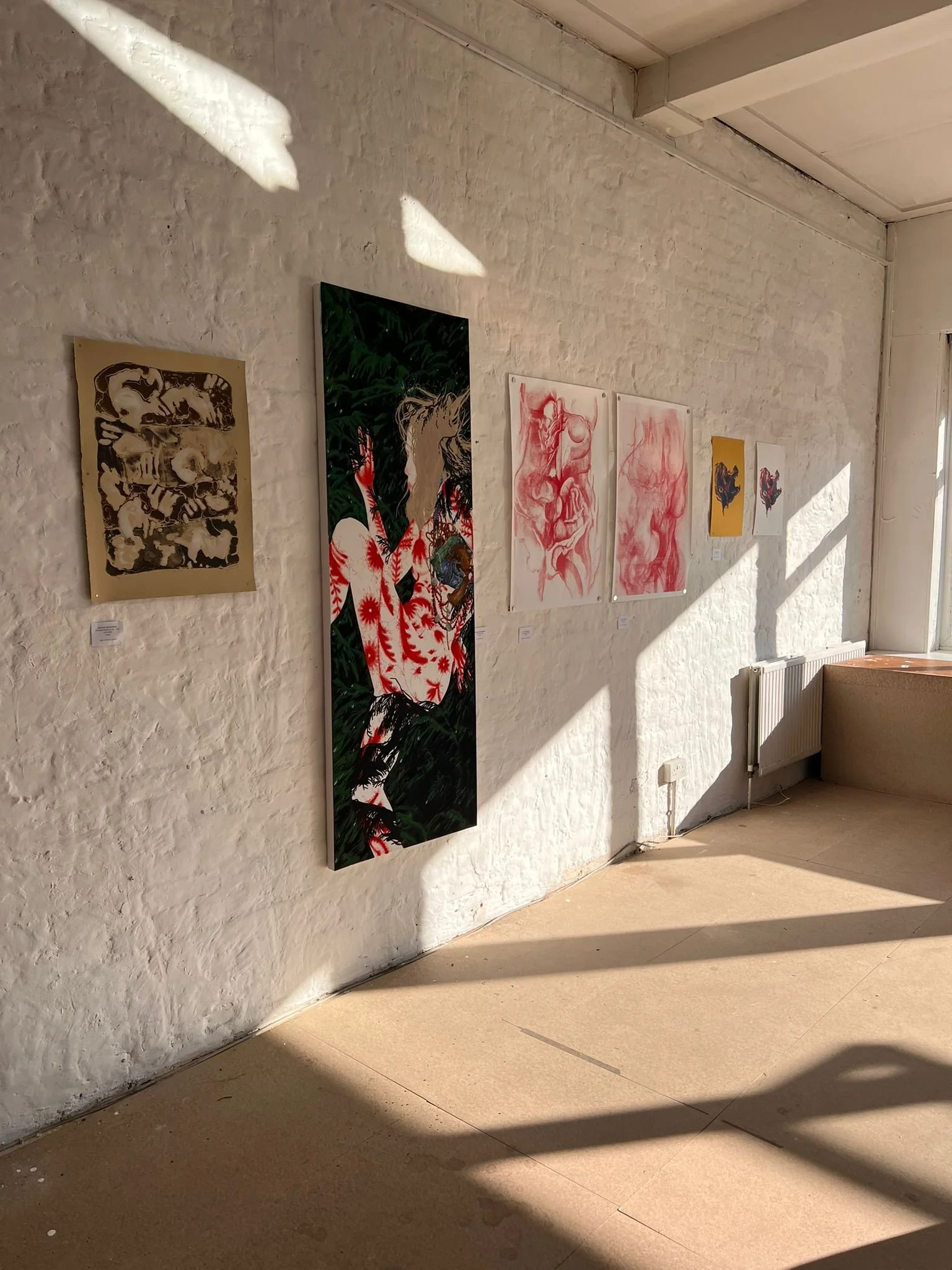 Gallery wall with seven paintings featuring abstract and figurative art, with a brick wall and sunlight casting shadows.