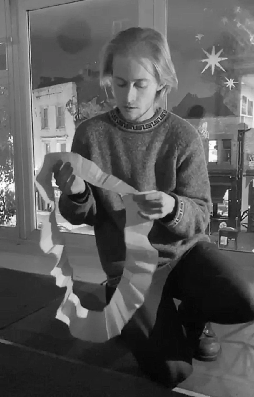 A young man with medium-length hair, sitting on the floor, is holding a large piece of paper with a torn paper ring or decoration, inside a room with large windows decorated with star and moon ornaments, and buildings visible outside.