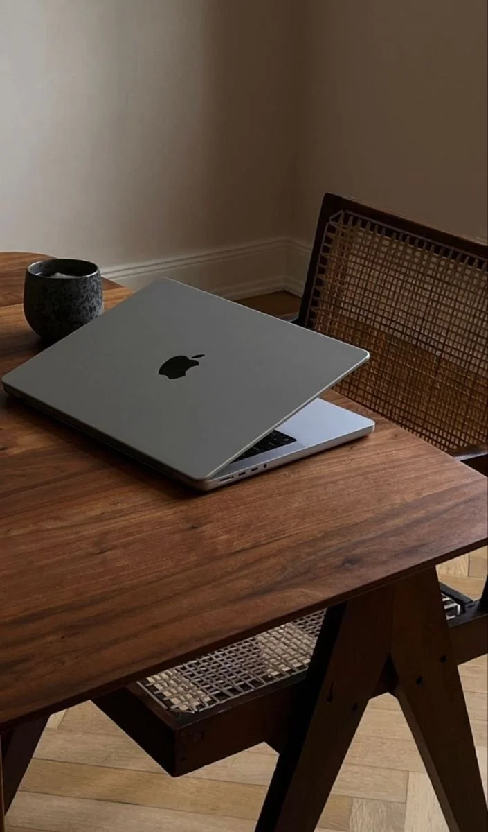 Wooden table with an open laptop, a coffee mug, and a woven chair.