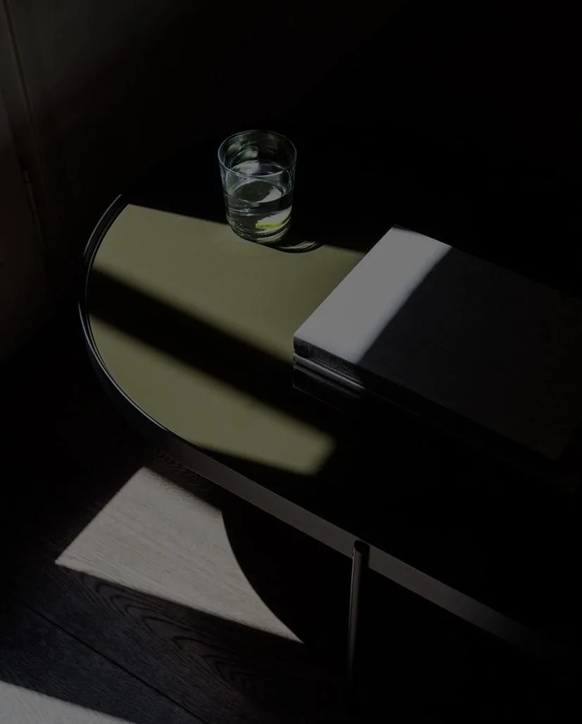 A glass of water on a round side table with a closed book on it, illuminated by sunlight creating shadows.