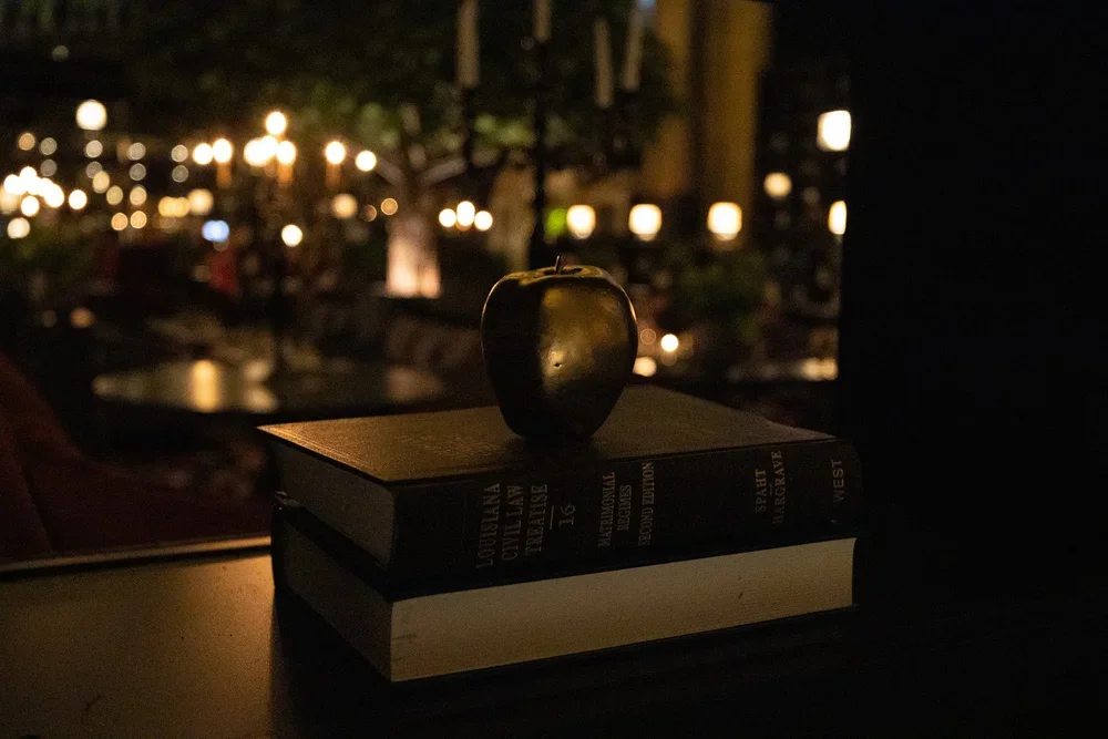 A black ceramic apple on top of two stacked books, in a dimly lit room with hanging lights and blurred background.
