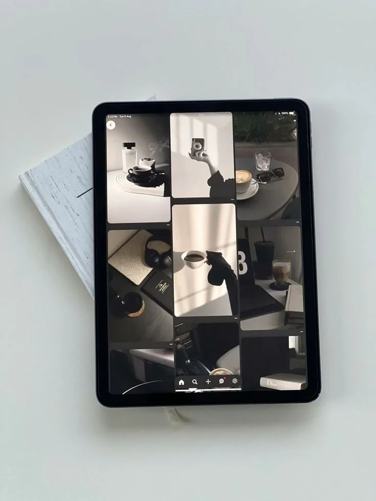 Tablet displaying a collage of nine black and white photos of coffee cups, a person holding a camera, headphones, and a car interior, placed on a white surface next to a closed notebook.